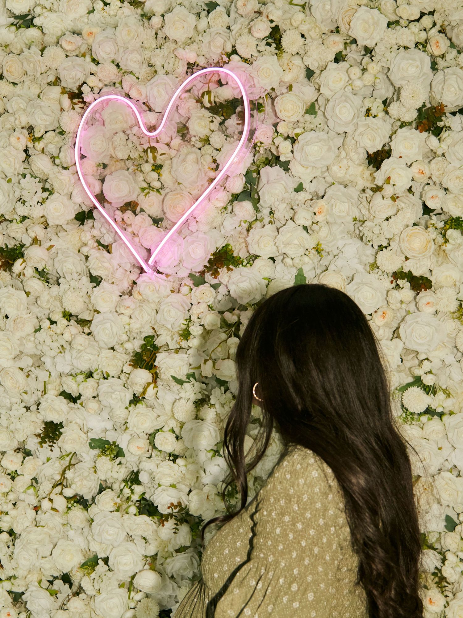 A maternity photo with pink neon heart sign against white flower wall backdrop.