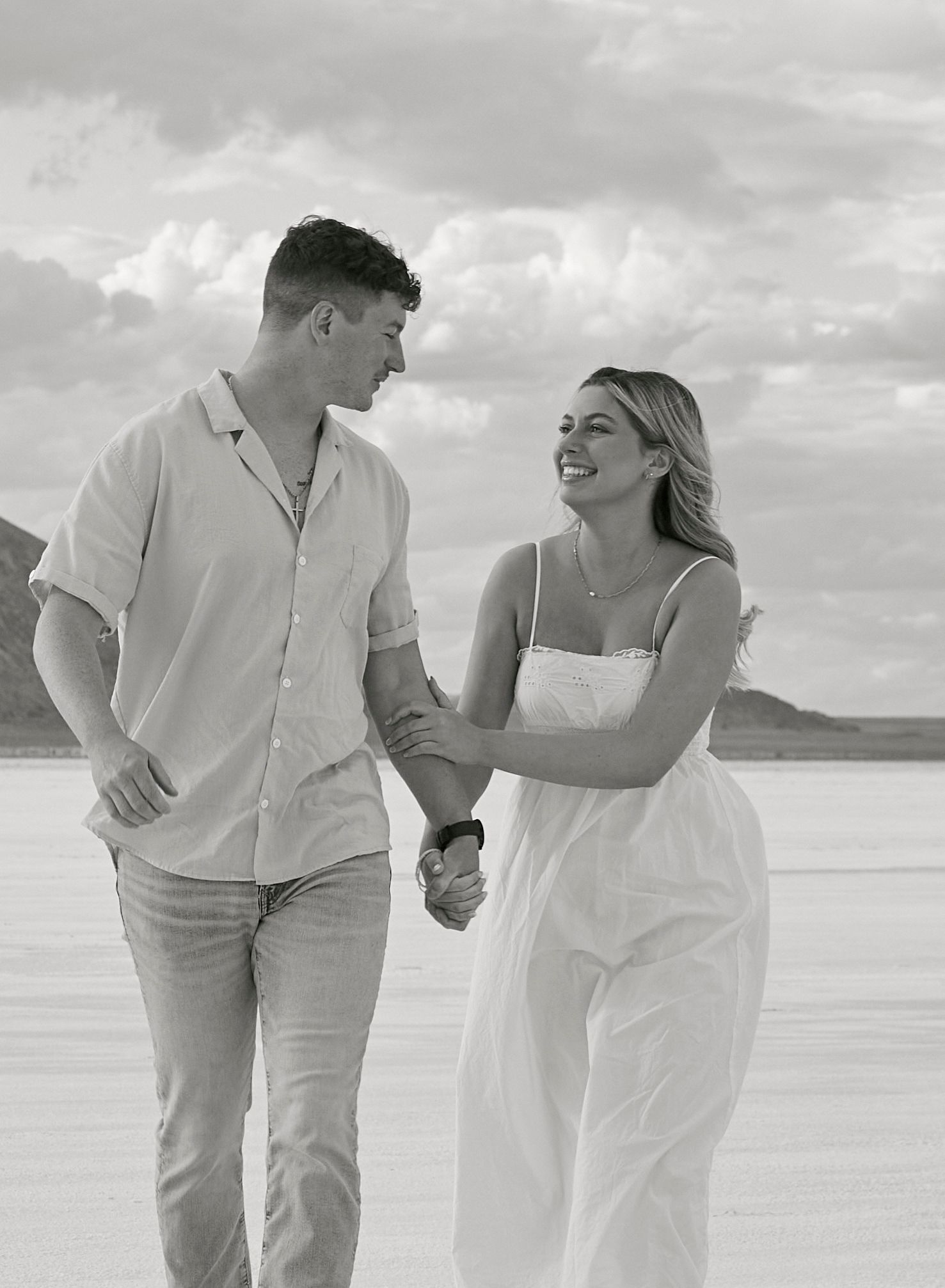 A couple walking hand in hand on a beach with mountains in the background.