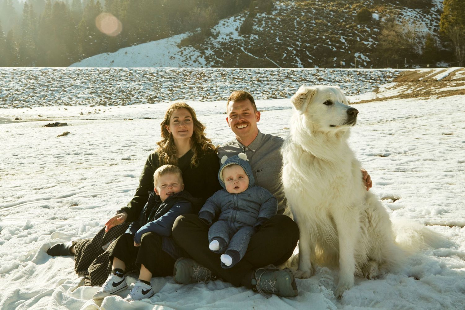 Family sitting together on snowy ground with their white dog in a winter mountain setting.