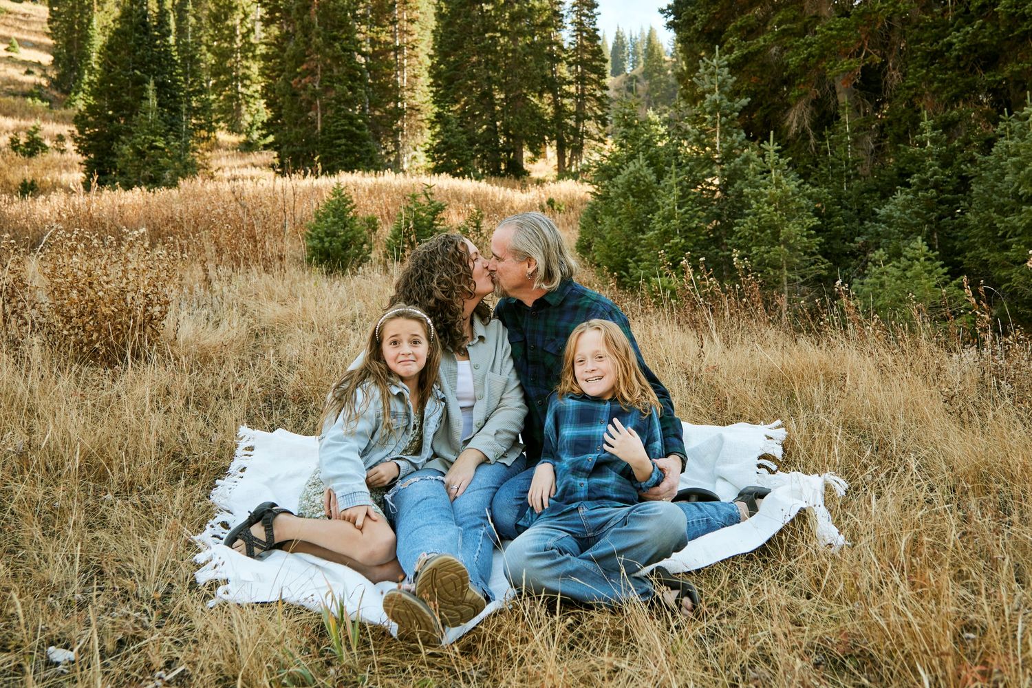 Family relaxing on white blanket in mountain meadow surrounded by pine trees.