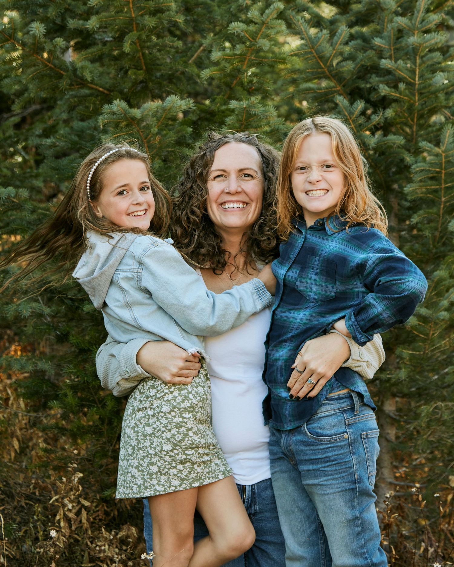 Family portrait among evergreen trees showing joyful embrace and casual style on sunny day.