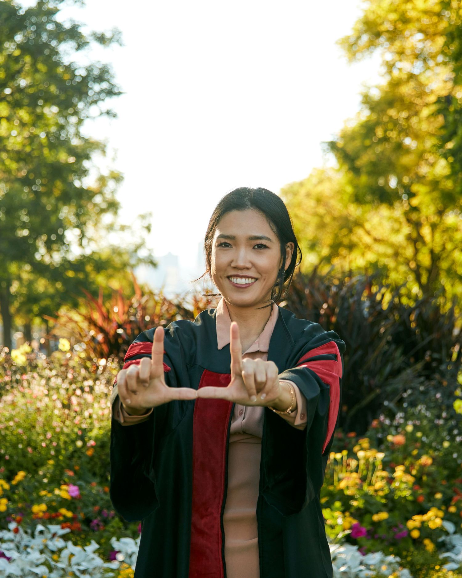 Person in graduation gown making heart gesture in garden setting.