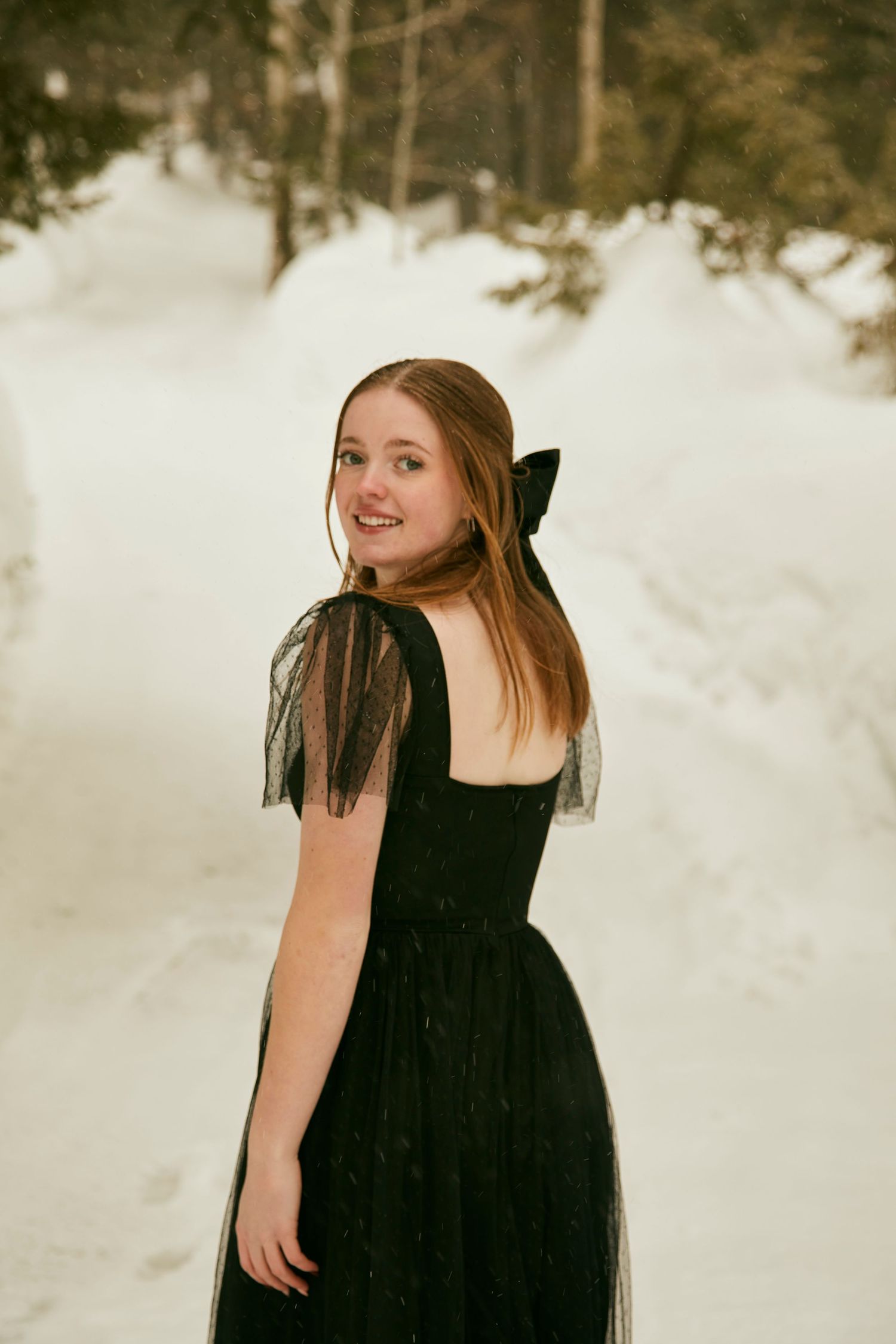 Person in black formal dress with tulle details poses in snowy winter setting.