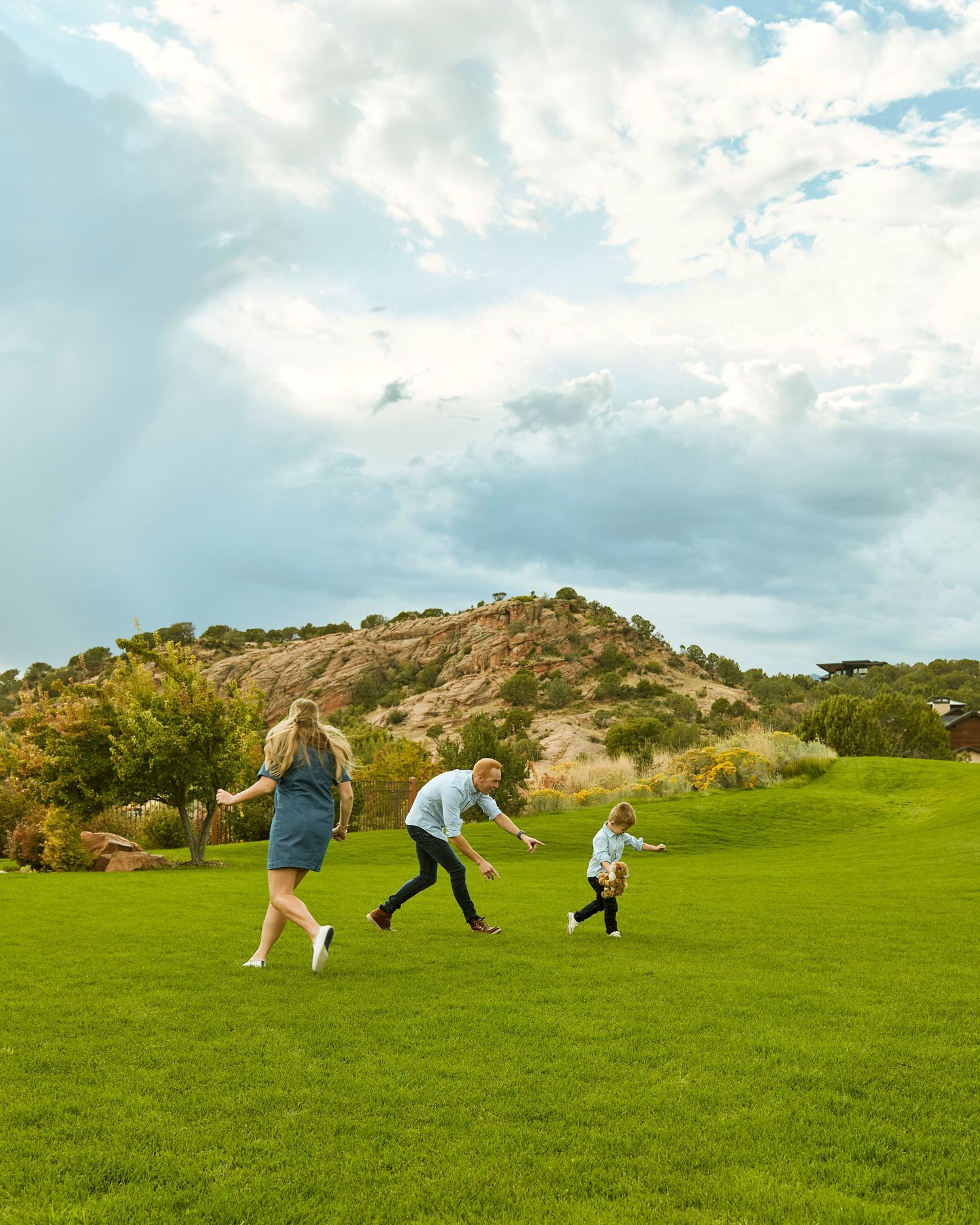 Three people running and playing on a green hillside under cloudy skies.