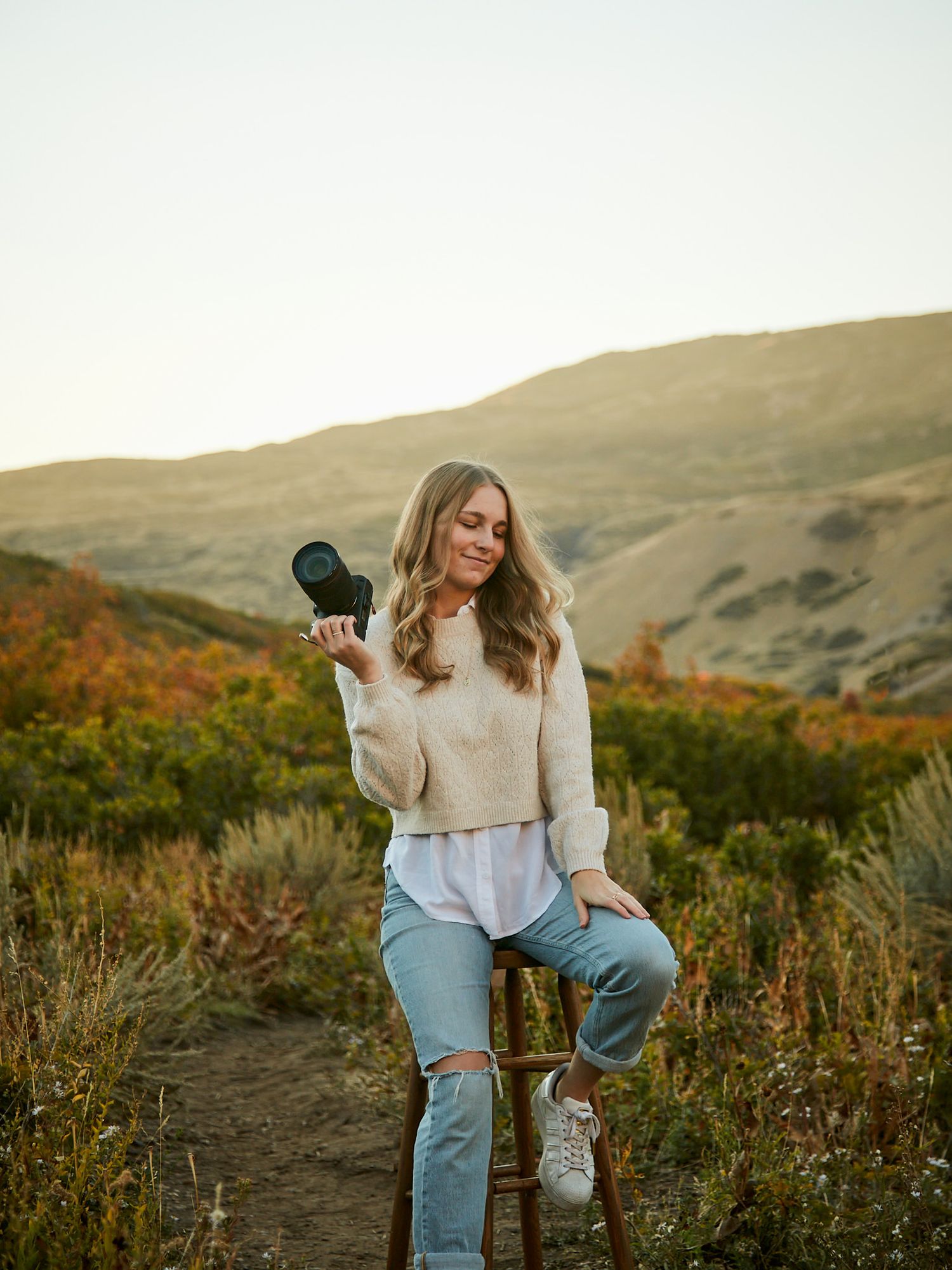 Casual portrait in cream sweater holding camera lens against autumn hillside backdrop.