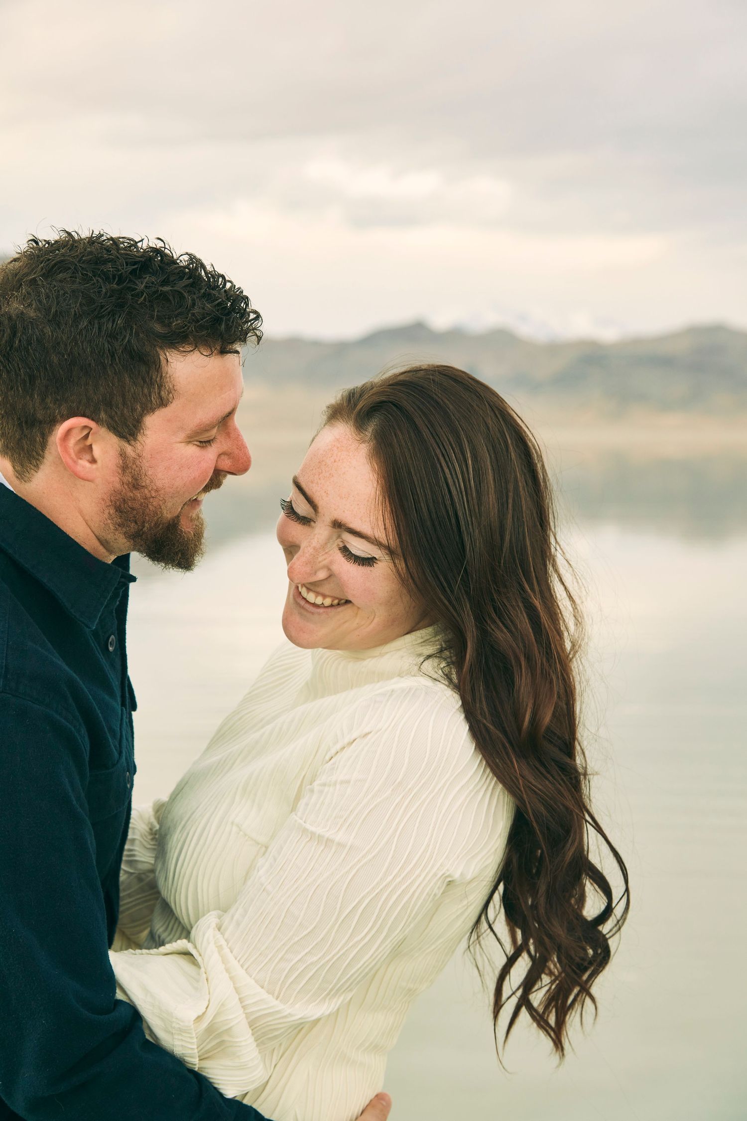Happy couple embracing by a serene lake with mountains in the background at sunset.