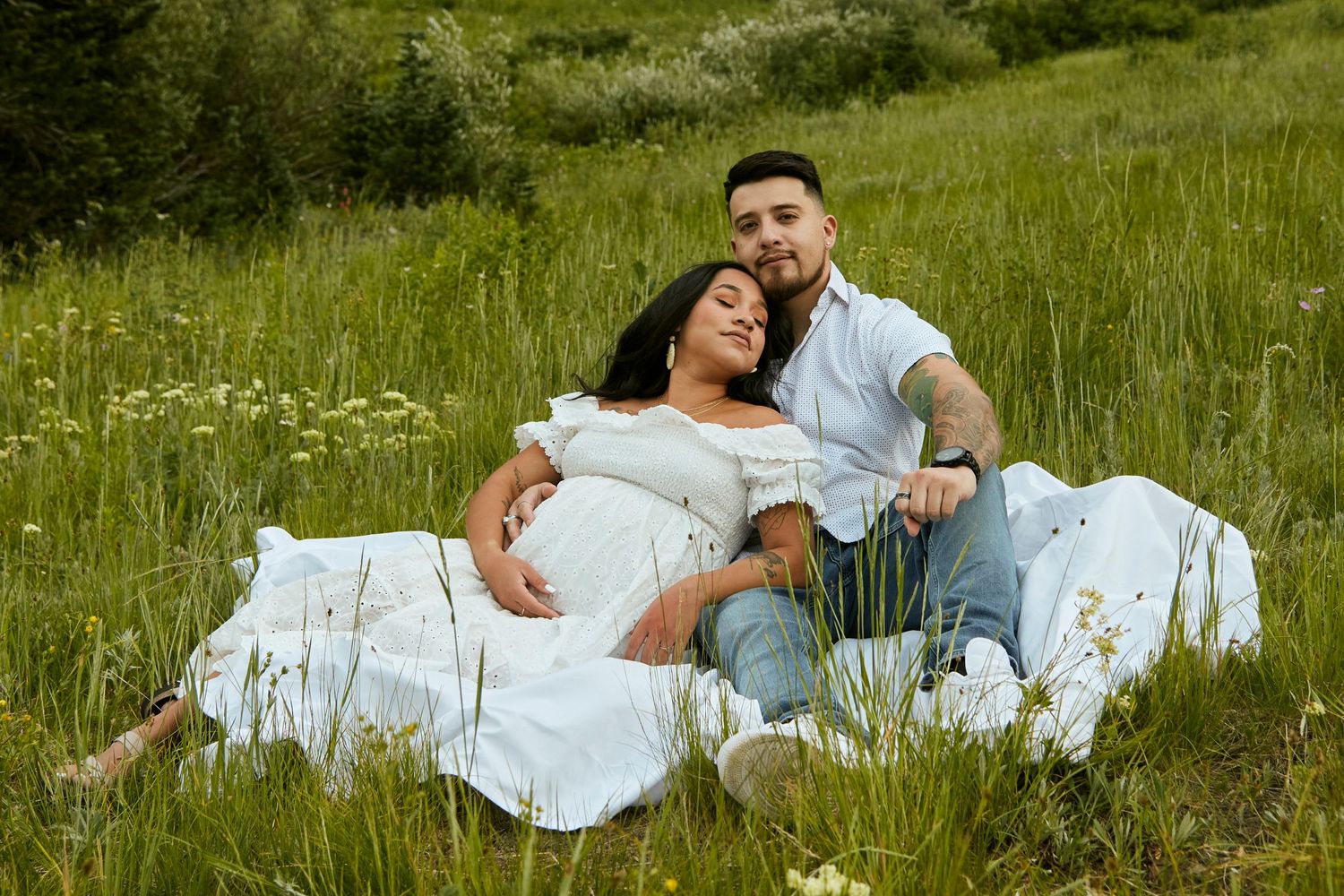 Couple relaxes together on white blanket in mountain meadow with wildflowers and greenery.