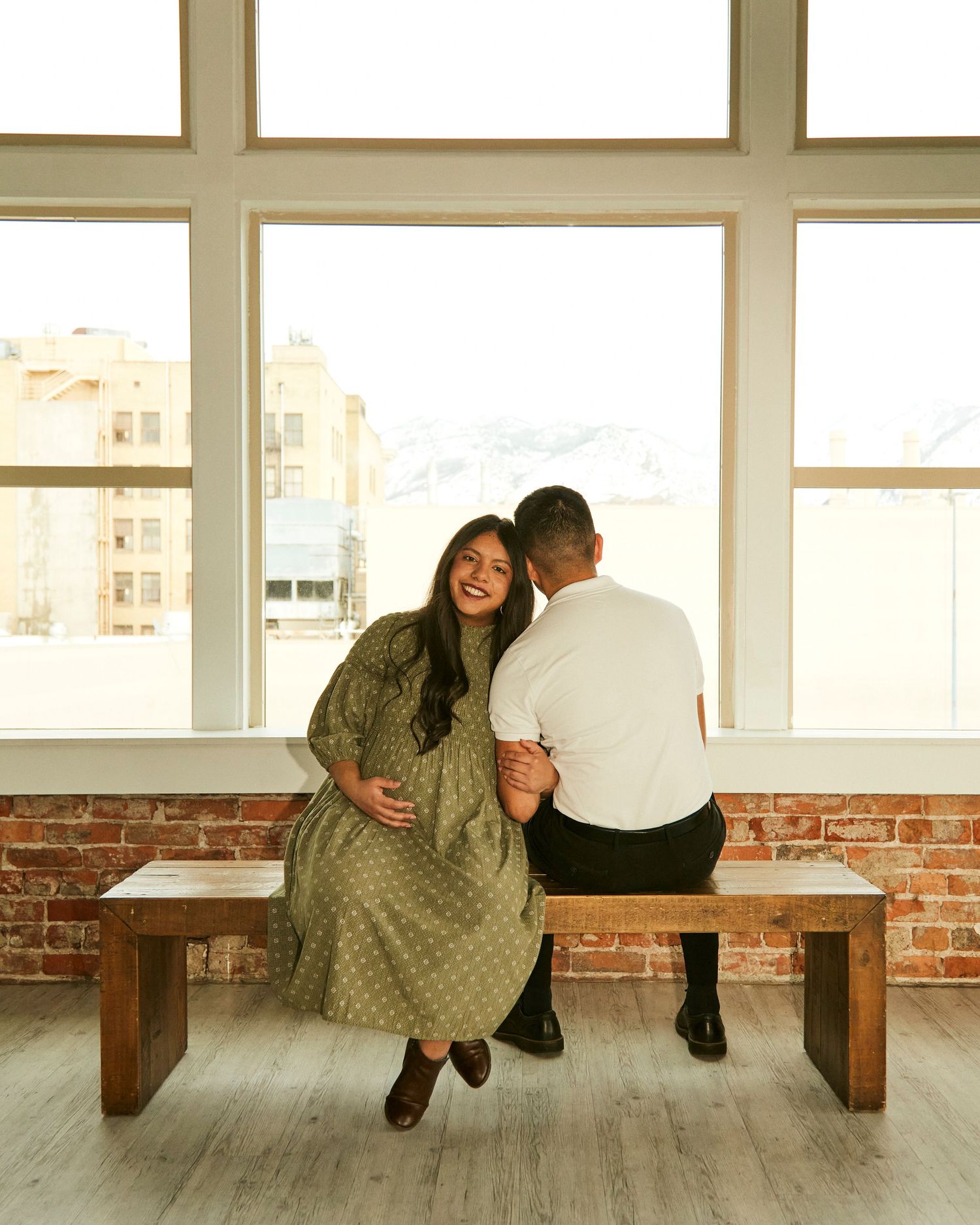 Couple sitting together on wooden bench by large windows with brick wall background.