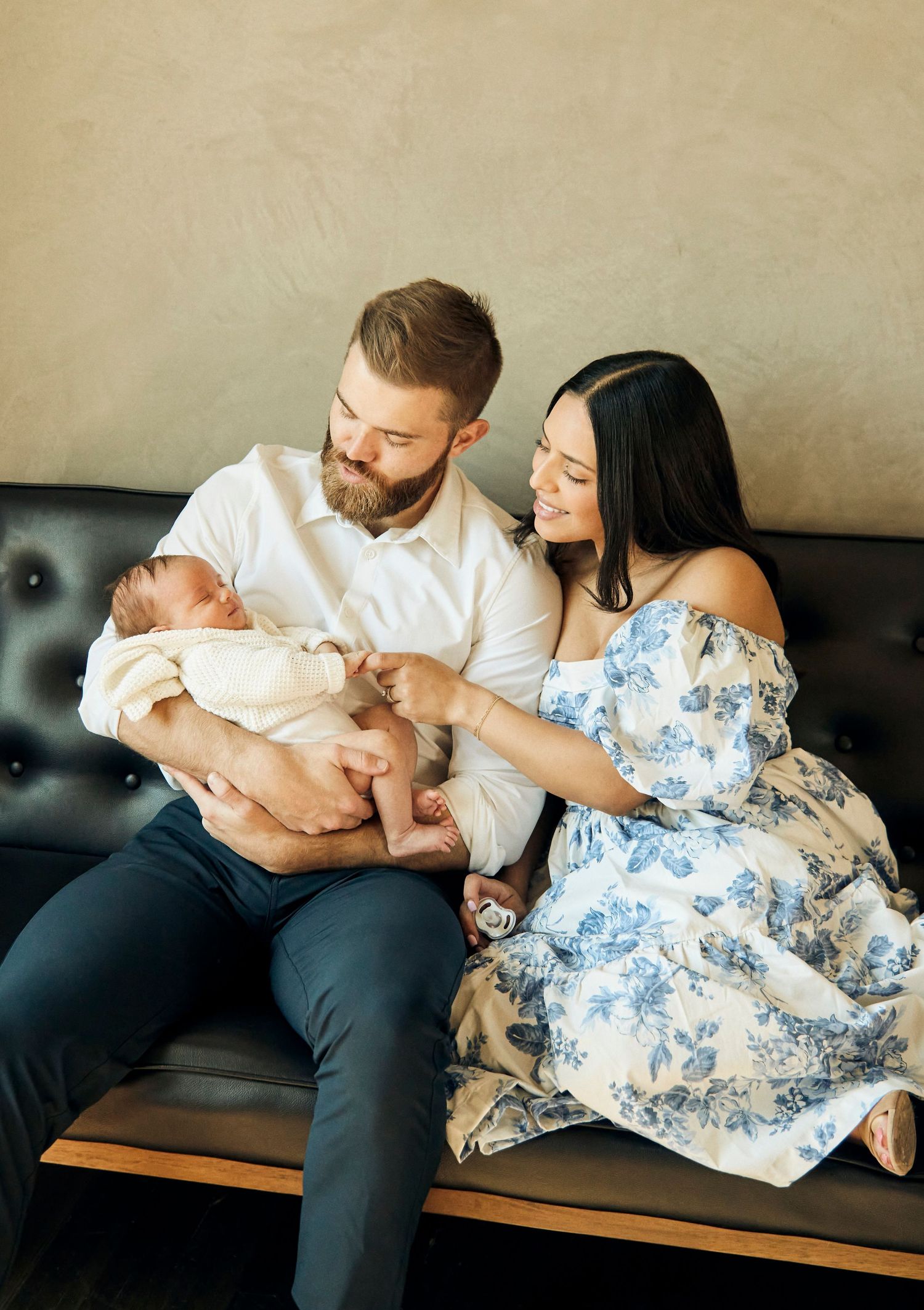 A couple sitting on a black leather couch admires their newborn baby while wearing casual attire.