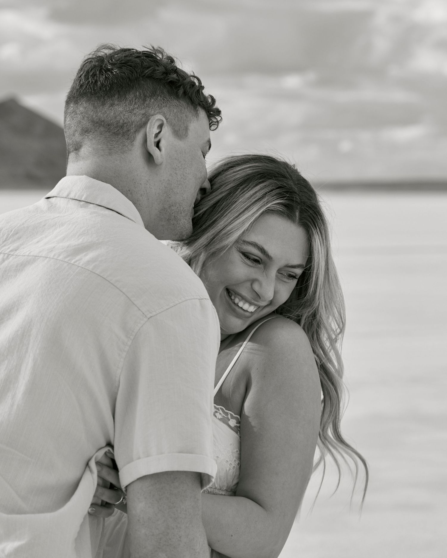 A black and white candid moment of laughter between couple on salt flats with mountains behind.