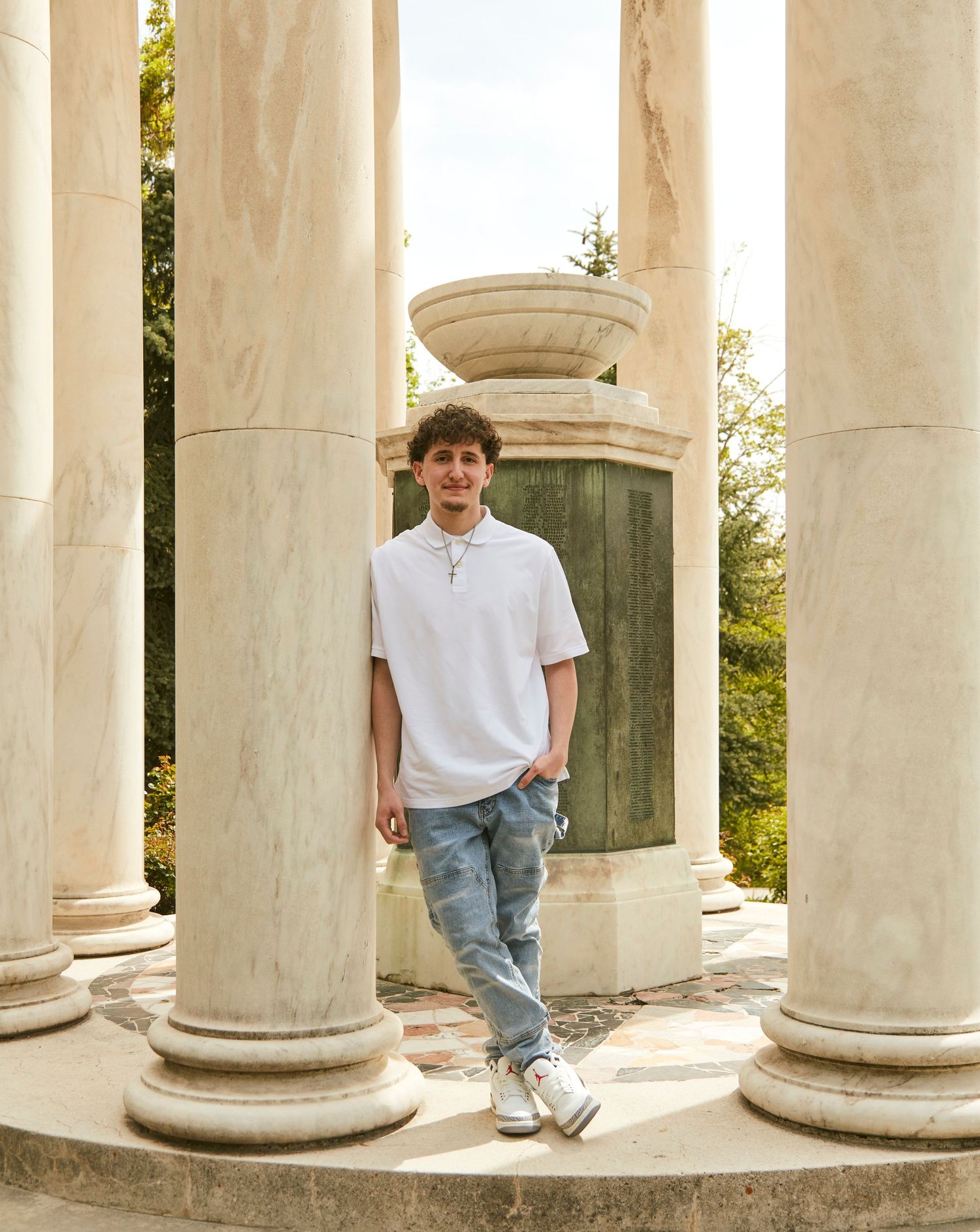 Young person in casual white polo and jeans poses between classical stone columns.