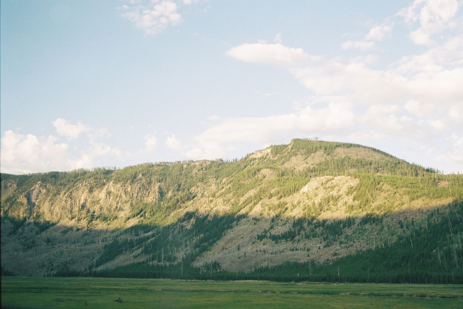 A rugged mountainside with exposed rocky cliffs rises above a green meadow under a partly cloudy sky.