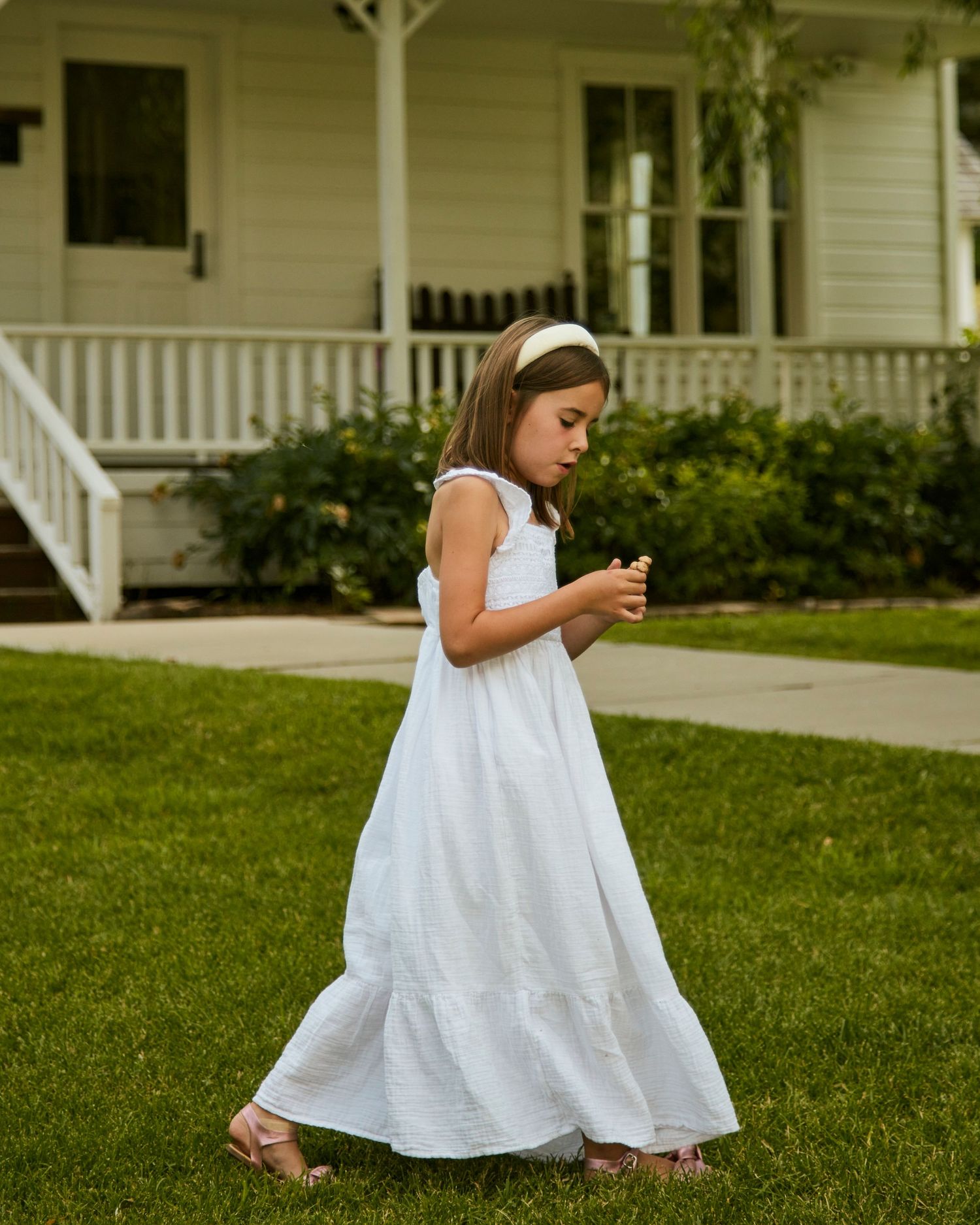 A young person in a white summer dress walks on grass in front of a classic white farmhouse porch.
