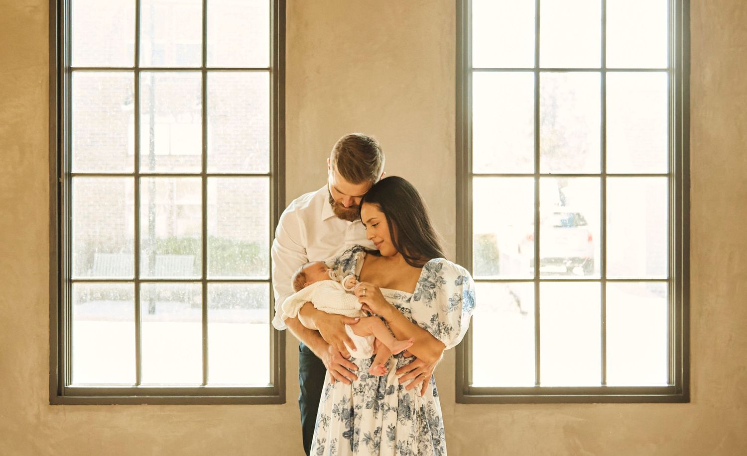 Parents share intimate moment with newborn baby by bright windows in rustic room.