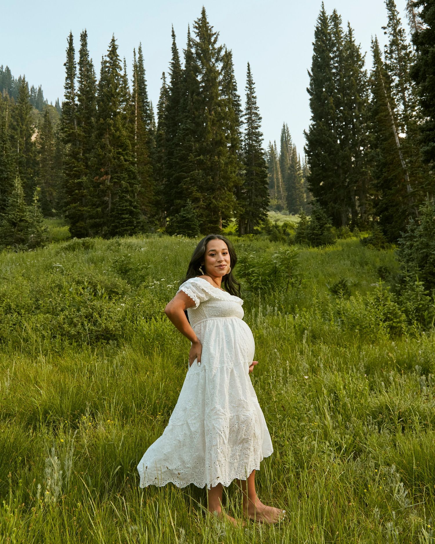A peaceful maternity portrait set against tall evergreen trees in a mountain meadow.