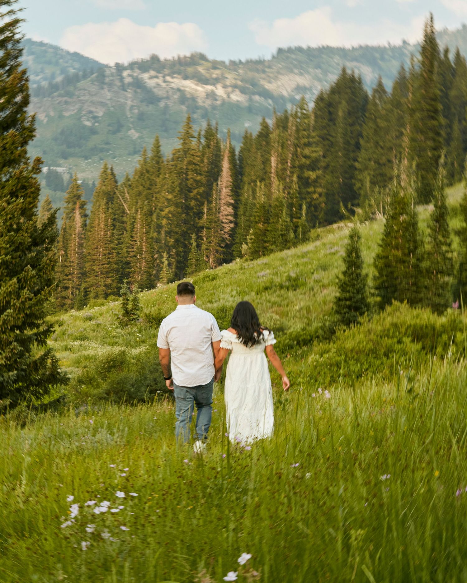 A couple walking hand in hand through a mountain meadow with evergreen forests and peaks behind them.