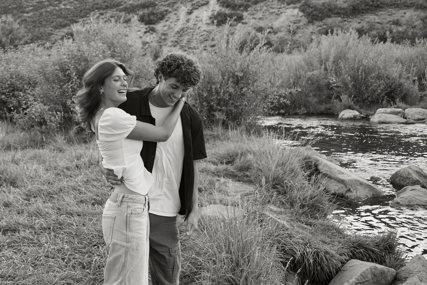 Romantic moment captured in black and white by a scenic river with rocks and grass.