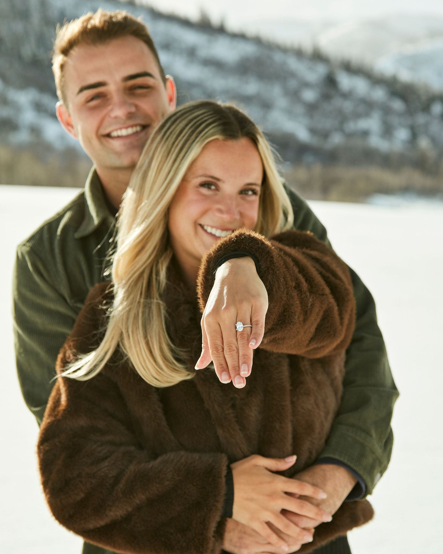 Newly engaged couple showing off ring in snowy mountain setting.