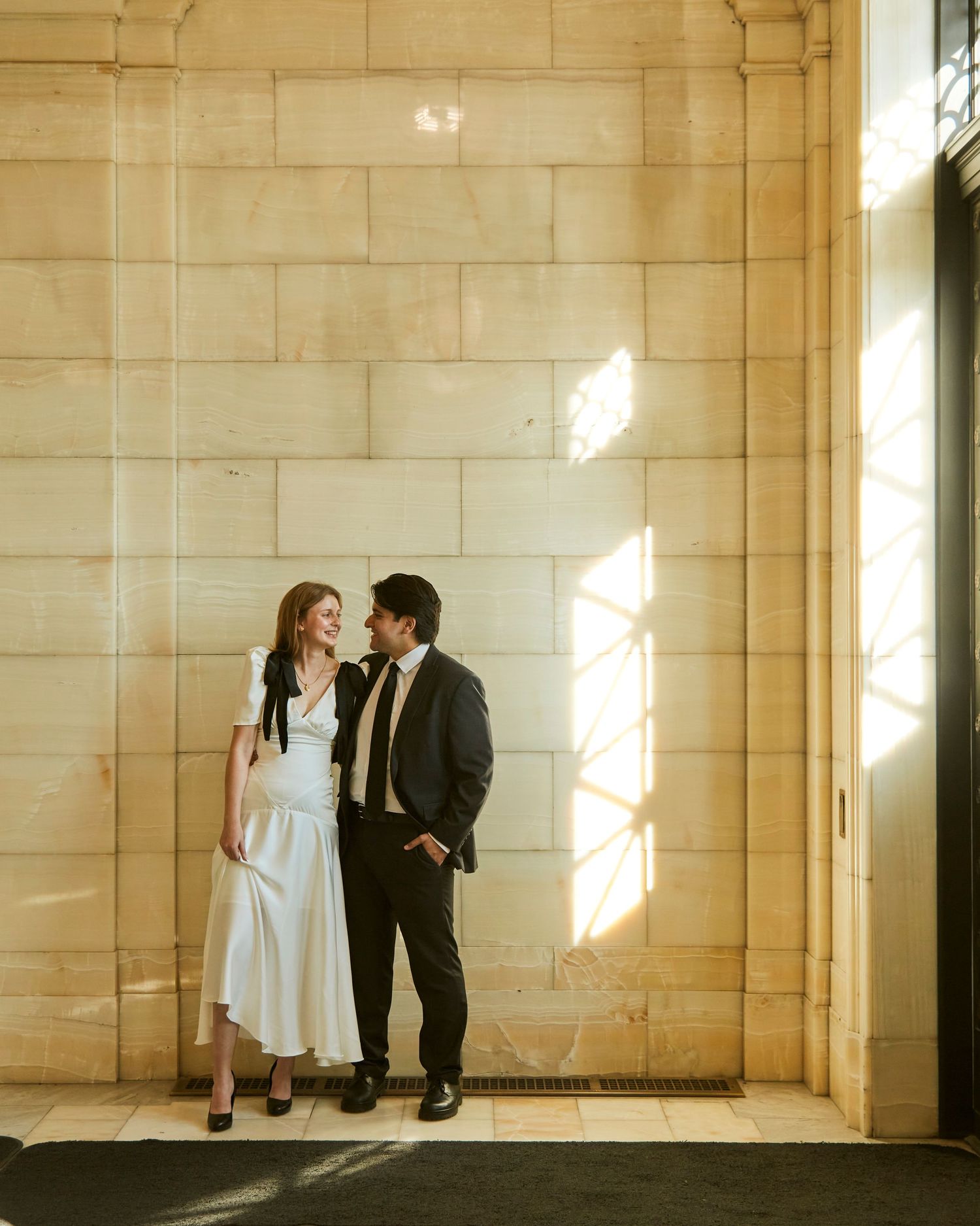 A couple poses against a sunlit stone wall in formal attire.