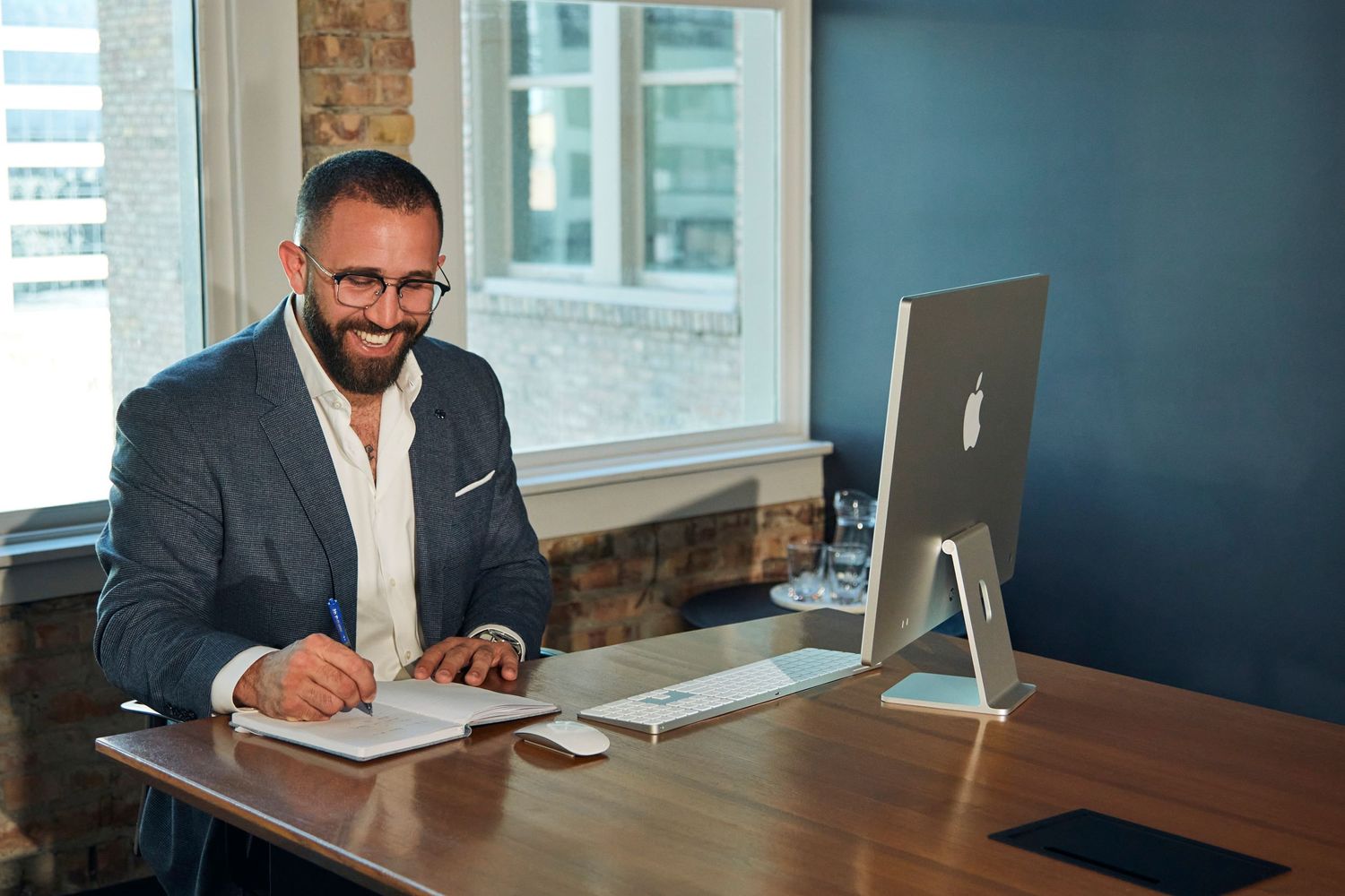 Business professional takes notes at modern workspace with iMac and exposed brick walls.