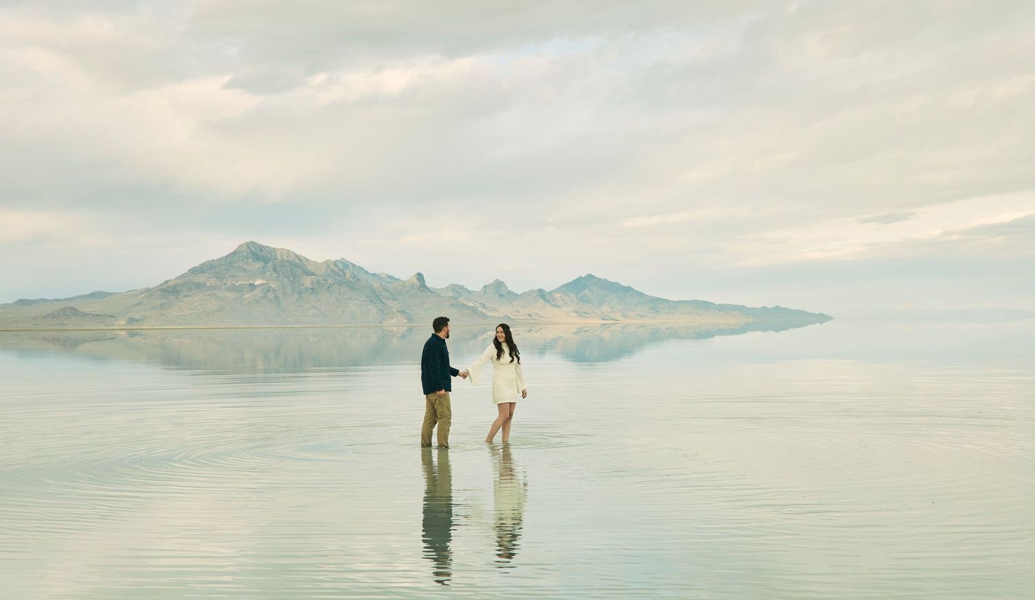 Couple standing in shallow water with mountains and cloudy sky in background.