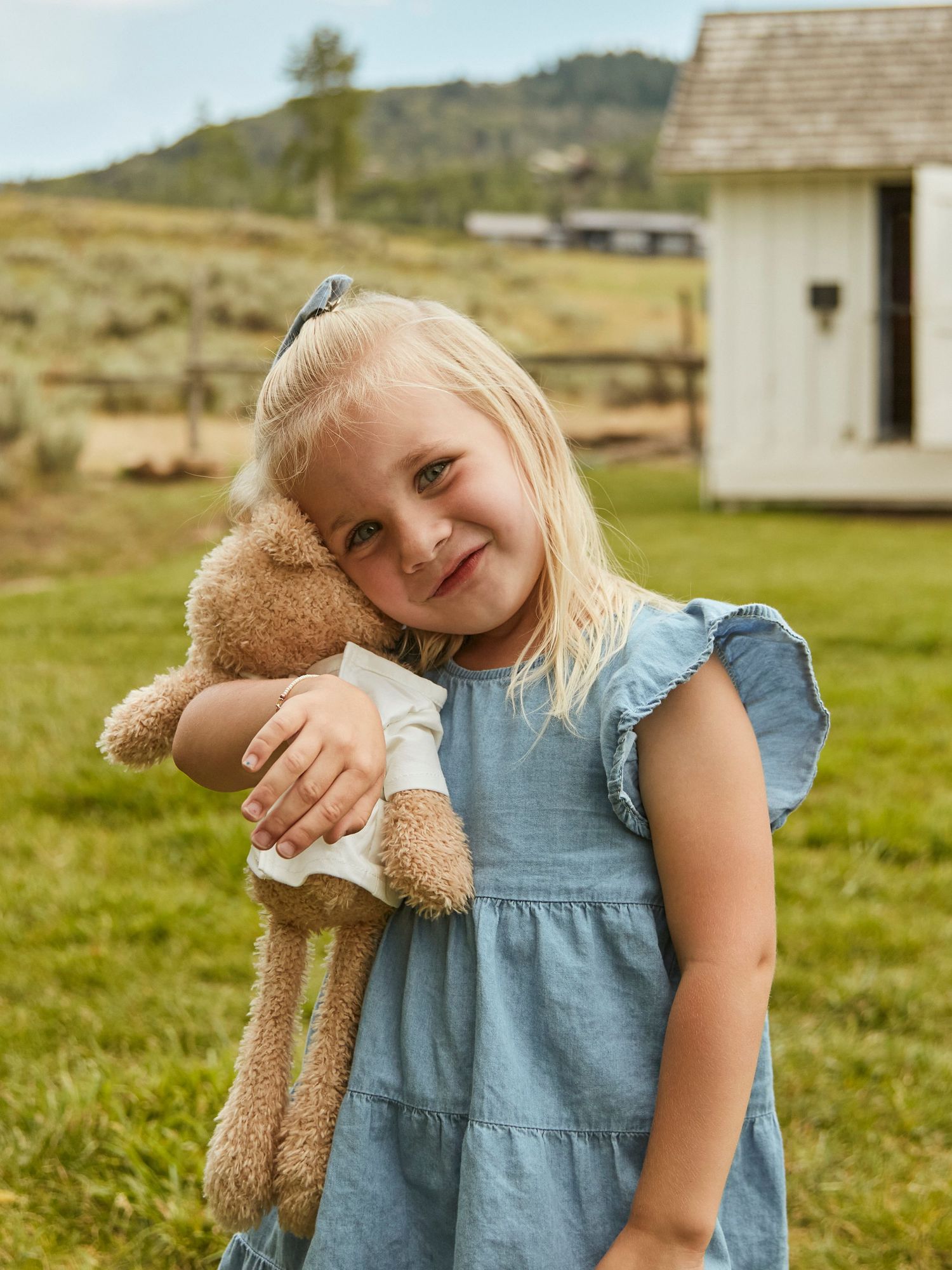 A child in a blue dress hugs a teddy bear while standing in a grassy field with a small building behind.