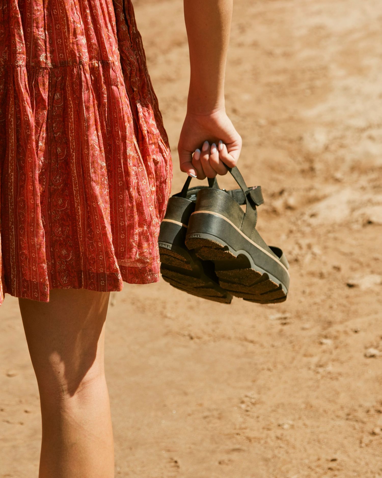 A person in a red dress holds a pair of chunky sandals while standing on sandy ground.