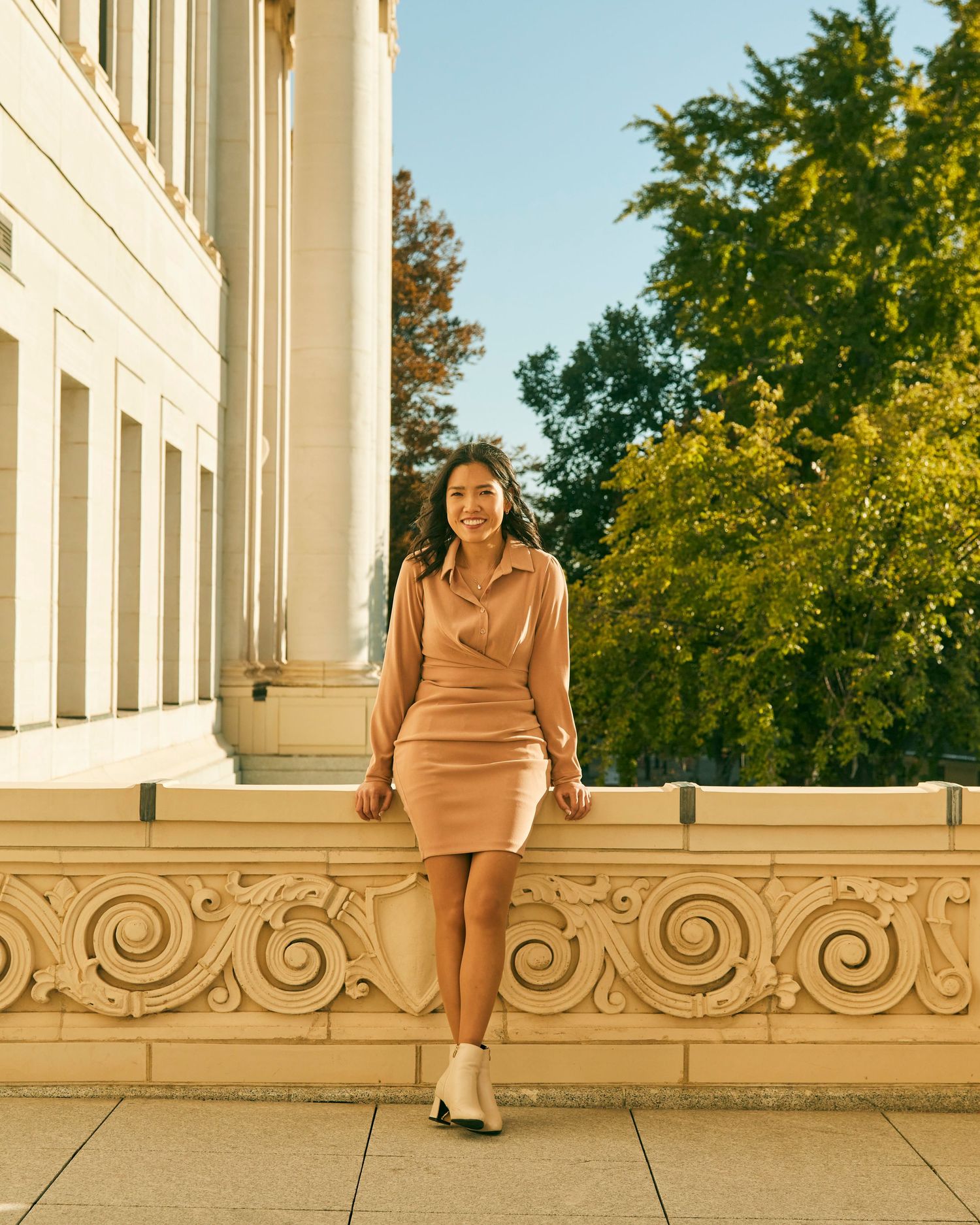 Woman in beige dress ensemble poses against classical architecture in warm sunlight.