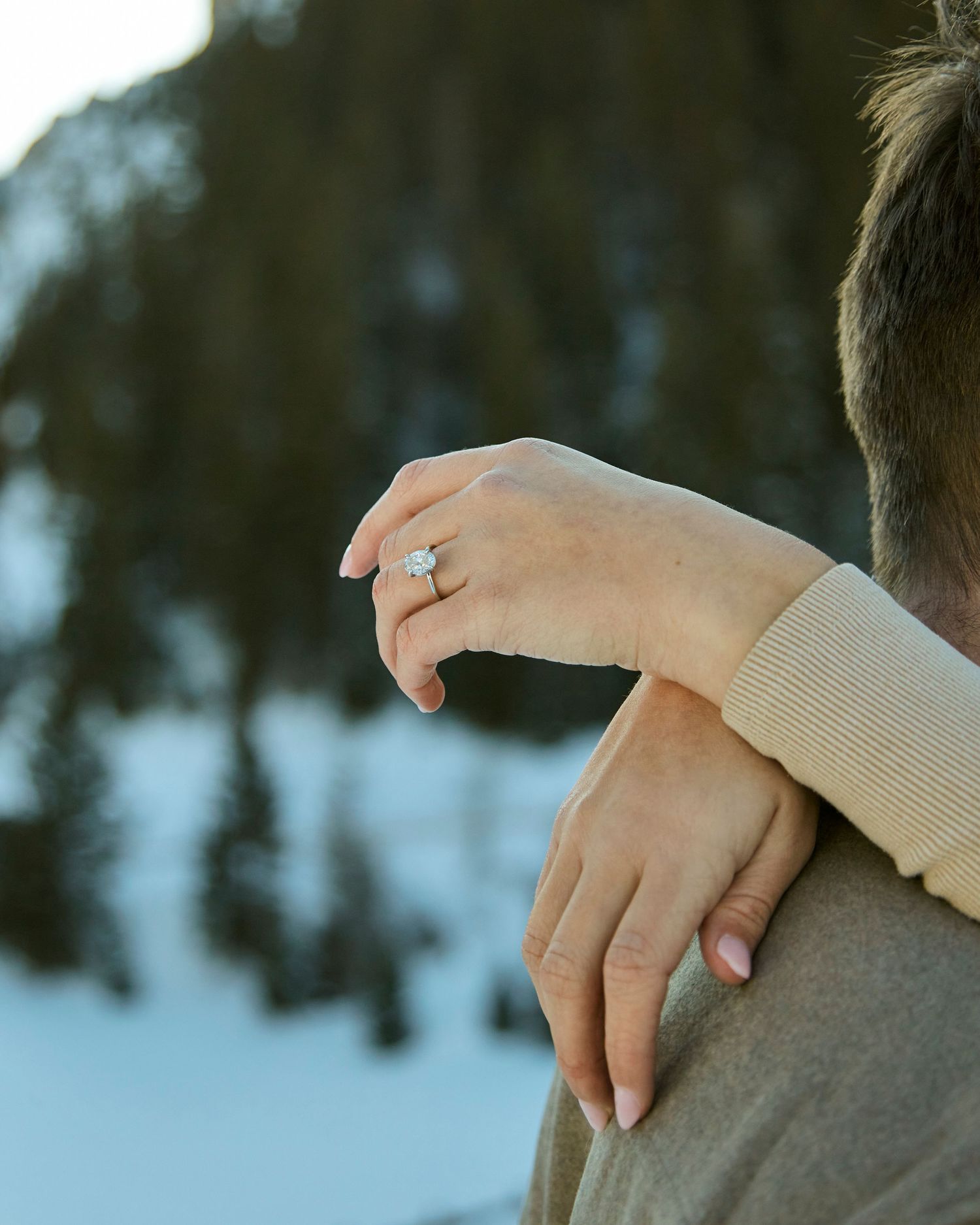 Close-up of hands with engagement ring embracing in snowy mountain setting.