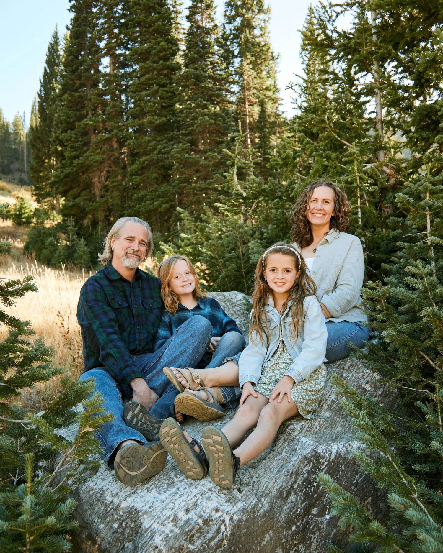 Family sitting together on a large rock surrounded by evergreen trees in a mountain setting.