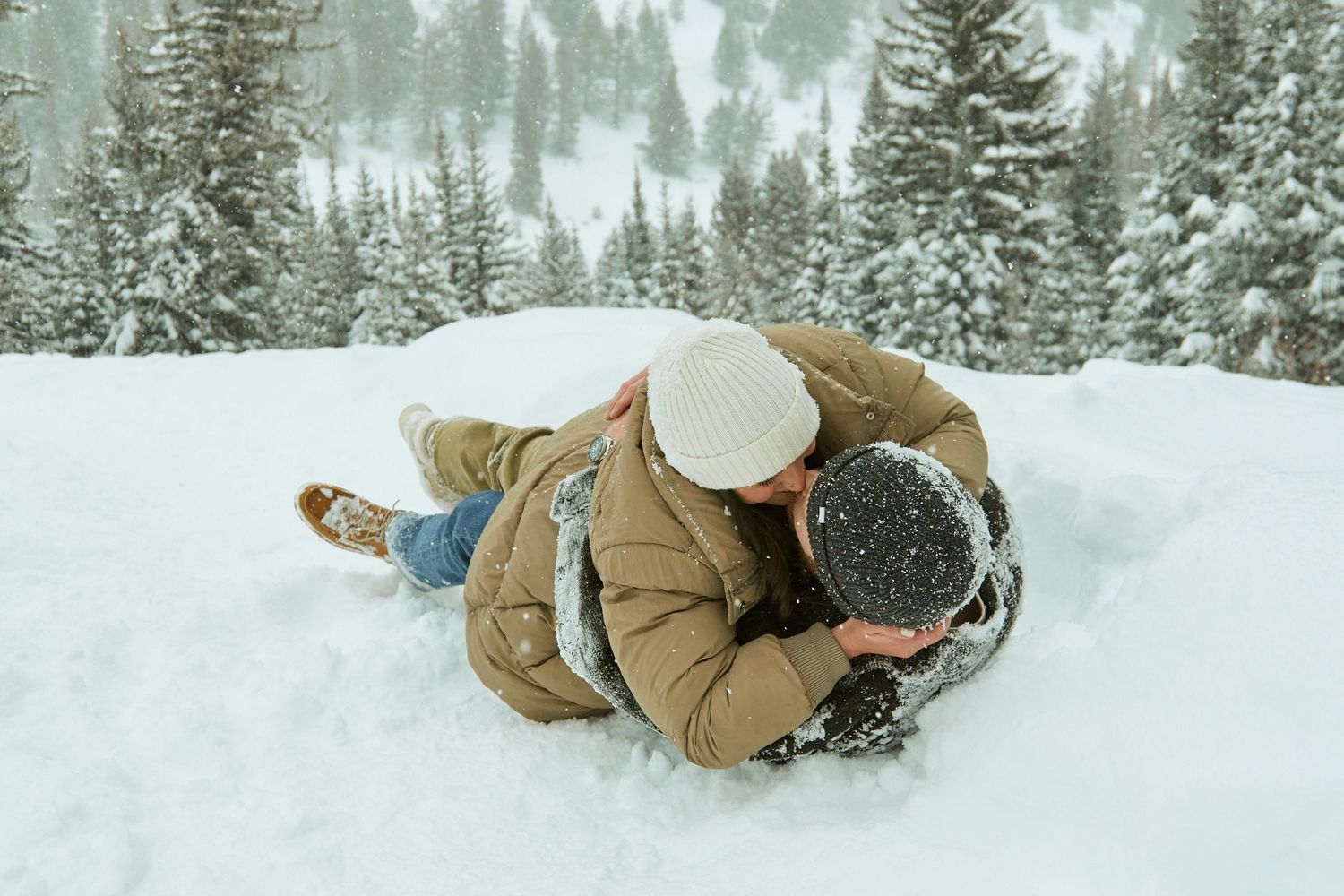 A couple plays in the snow wearing cozy winter coats during a romantic winter photo session in a snowy forest.