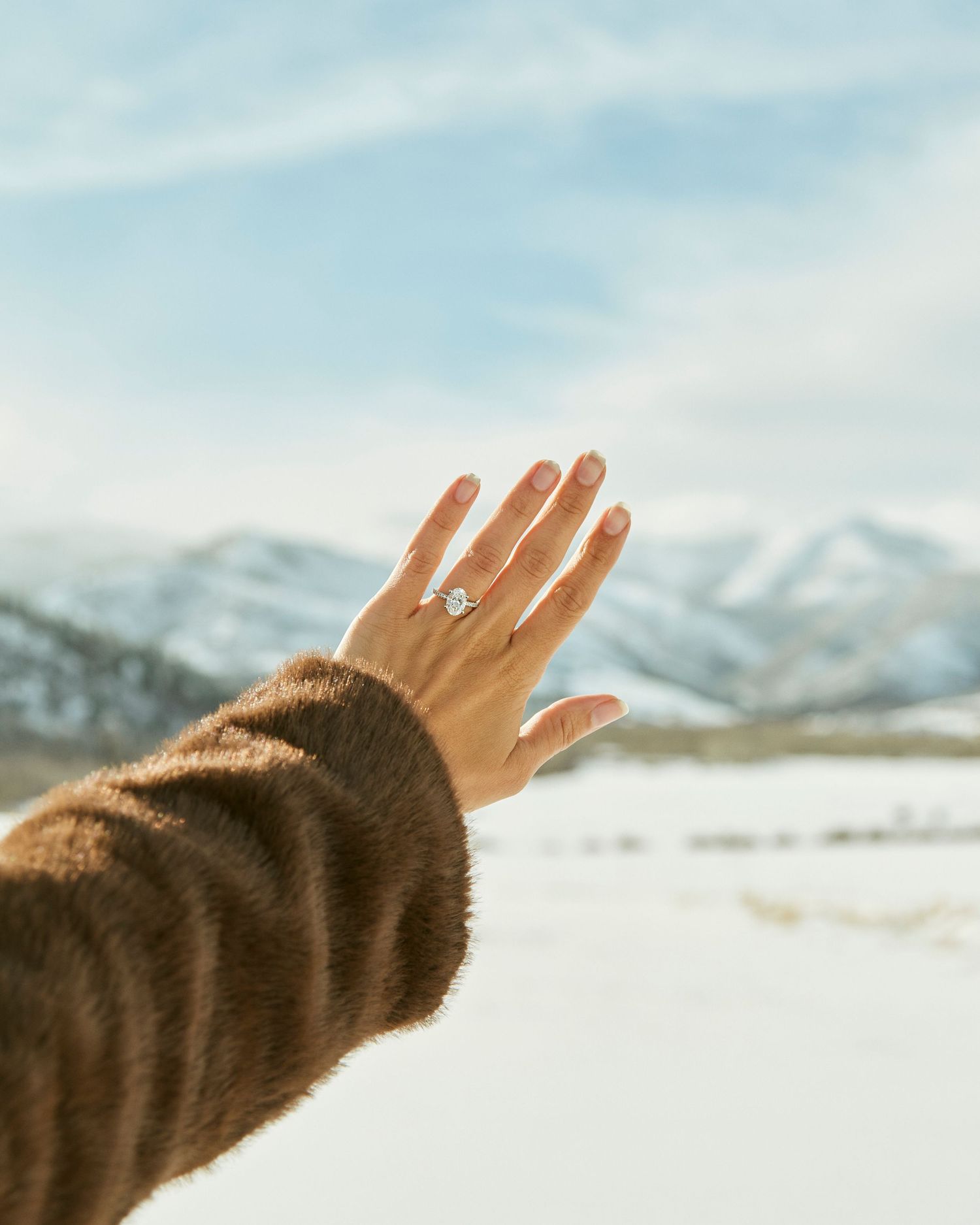 A hand wearing an engagement ring extends toward snow-capped mountains under a bright winter sky.