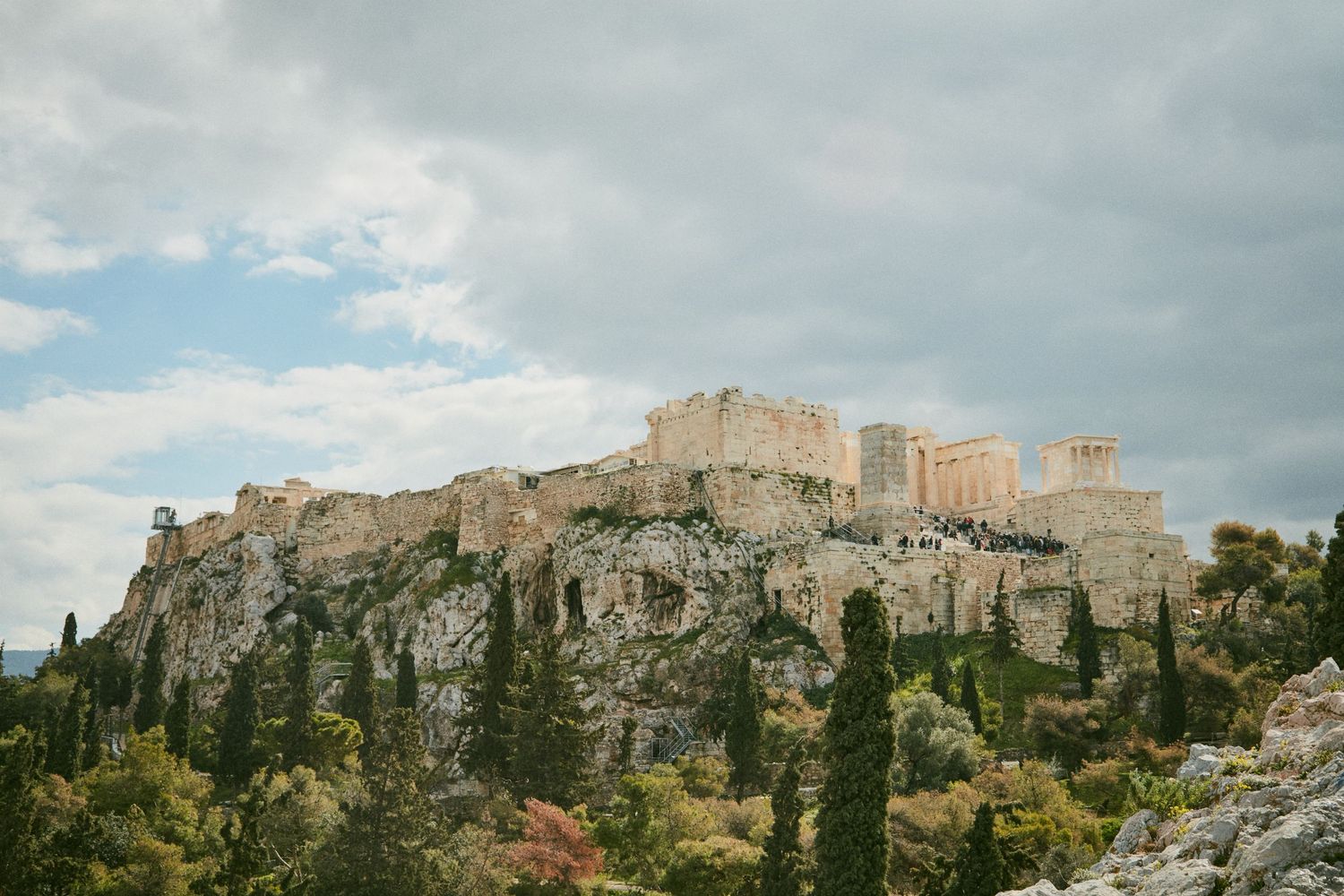 Ancient Acropolis of Athens towering over cypress trees against cloudy sky.