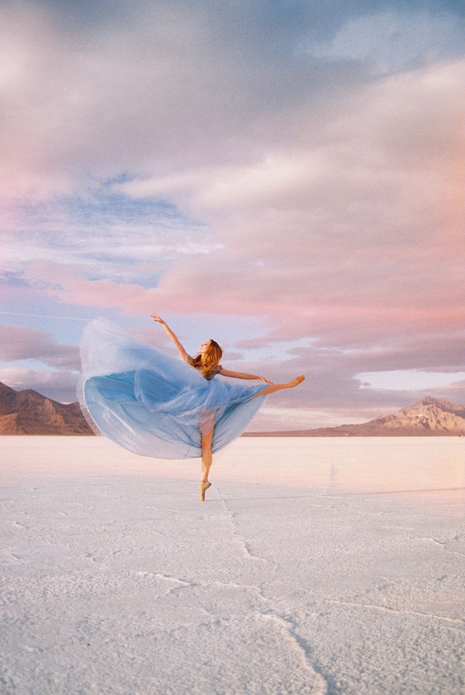 Ballet dancer poses gracefully in a light blue dress on white salt flats during sunset.
