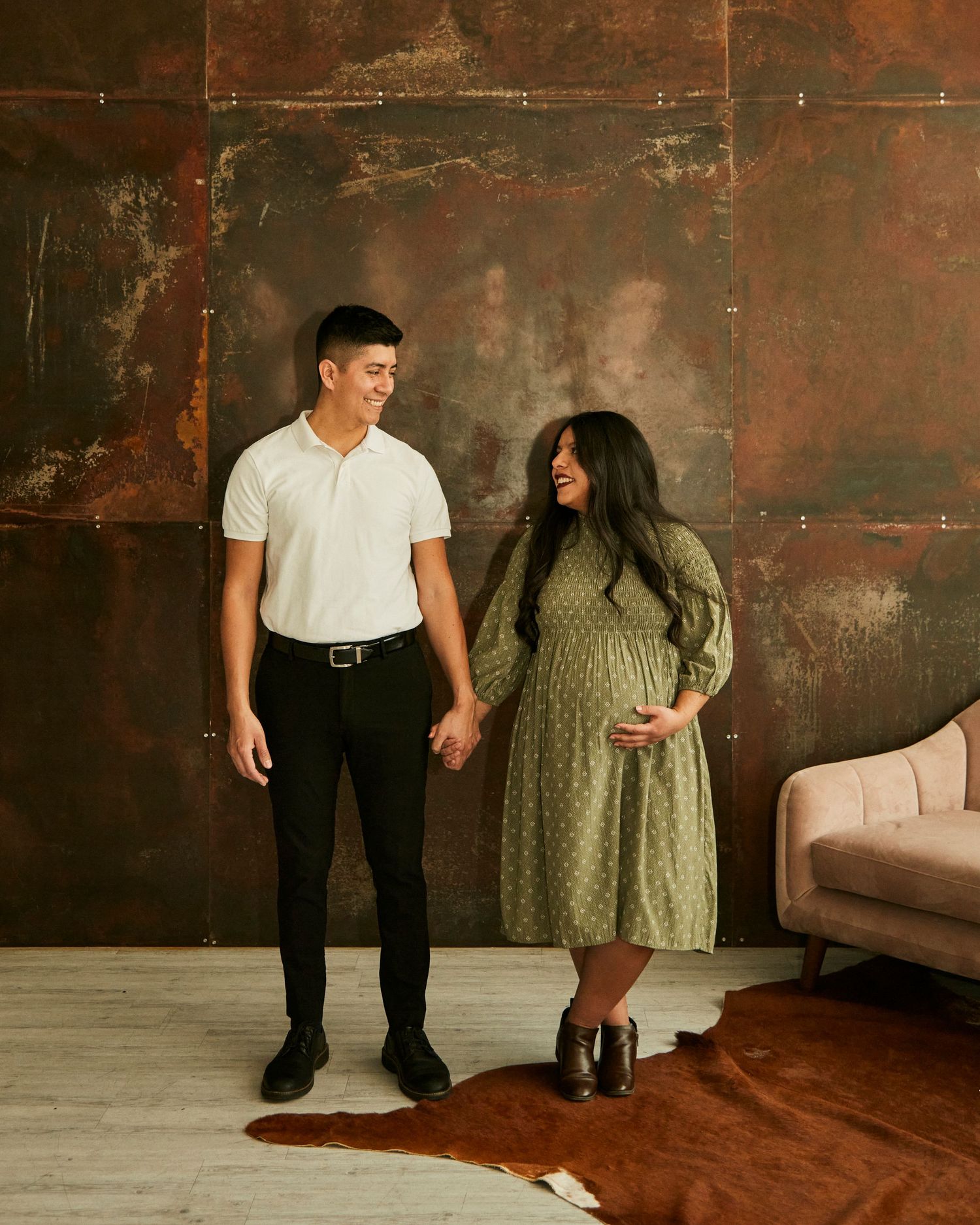 Couple stands holding hands against industrial metal wall with person in green polka dot dress.