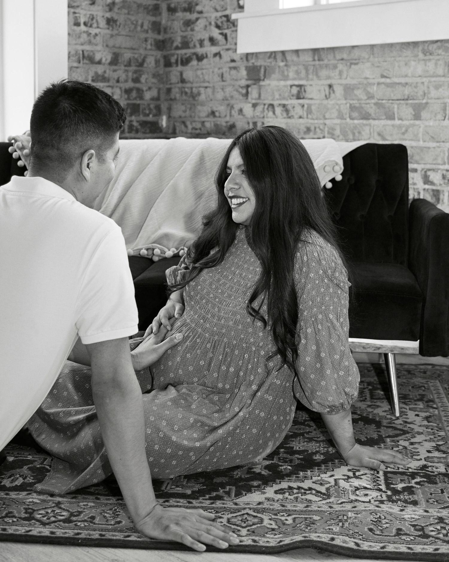 Couple shares a joyful moment on carpet with brick wall backdrop in black and white photo.
