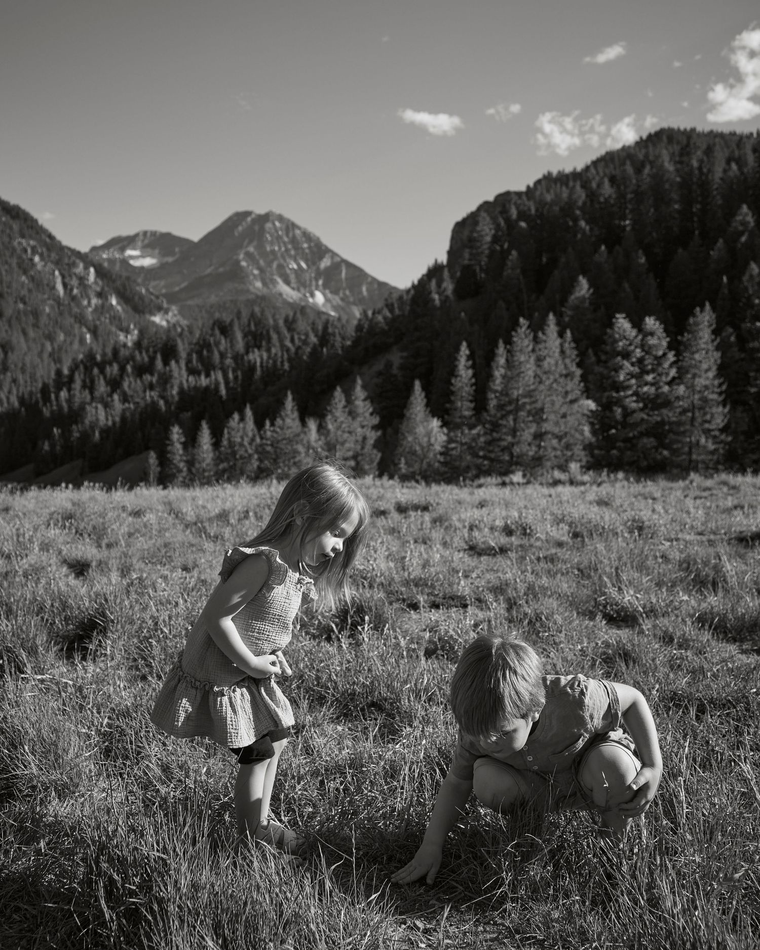 Children playing in a mountain meadow with dramatic peaks and forest in black and white.