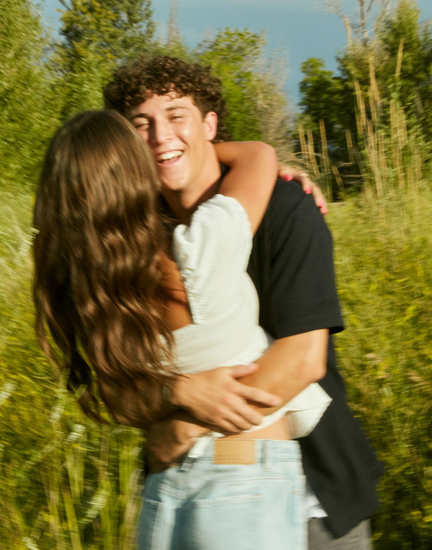 Couple sharing joyful embrace in sunlit field with trees in background.