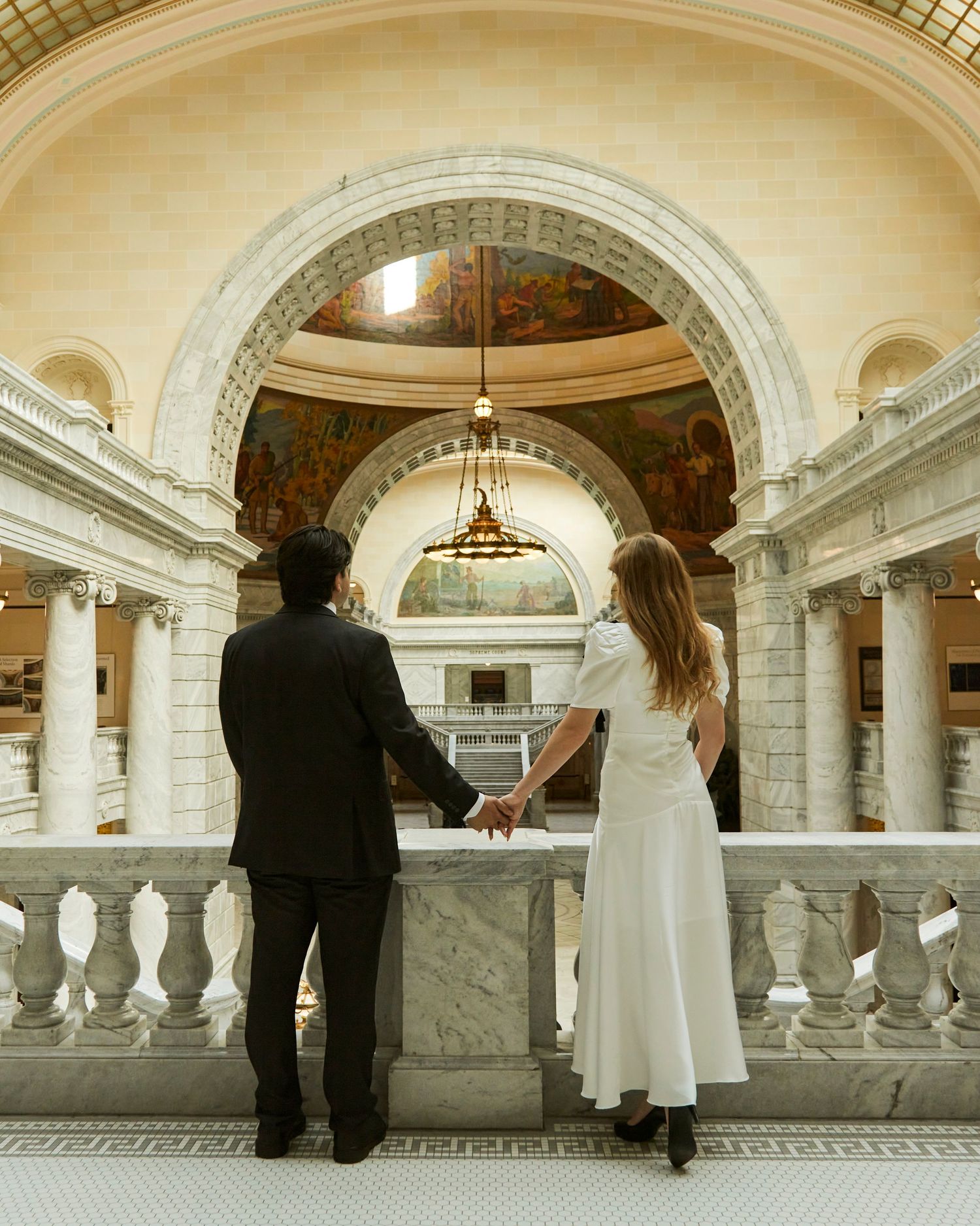 A couple stands at a marble balustrade overlooking an ornate domed interior.