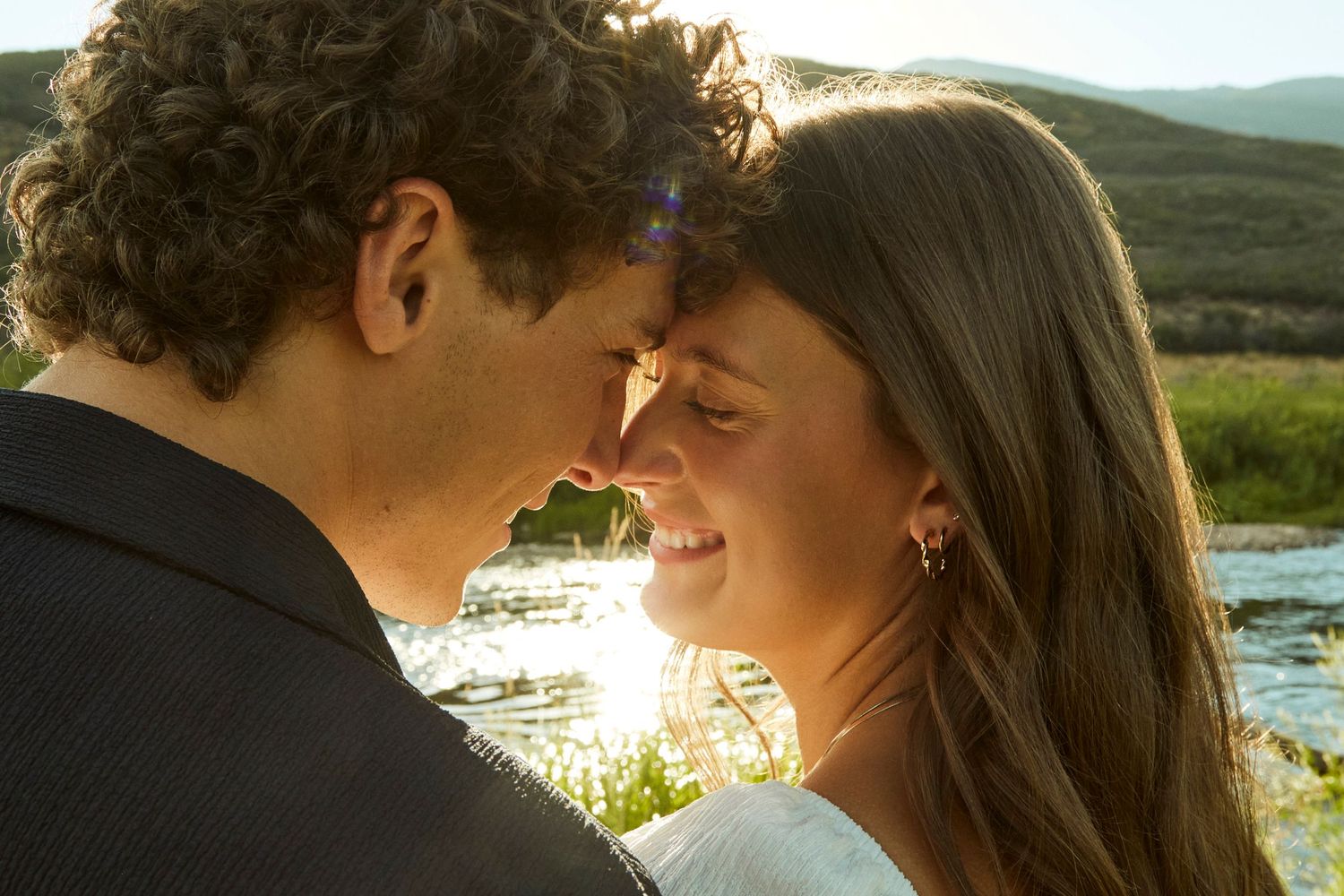 Intimate moment between couple at sunset by riverside with mountains in background.