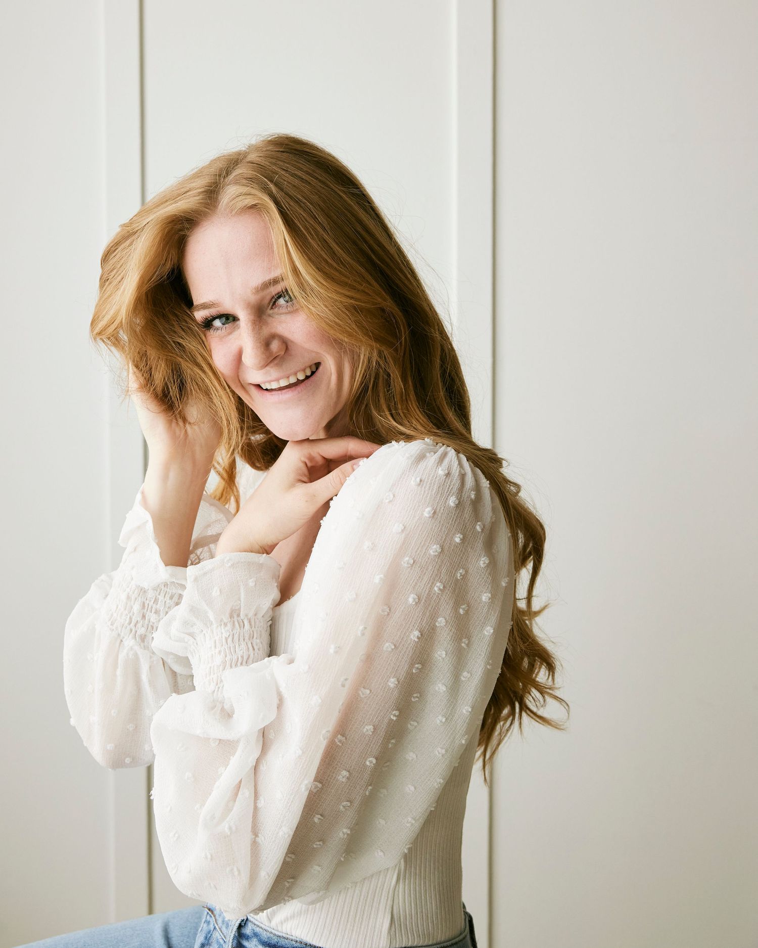 Person in white sweater with long hair smiles joyfully against light wall.