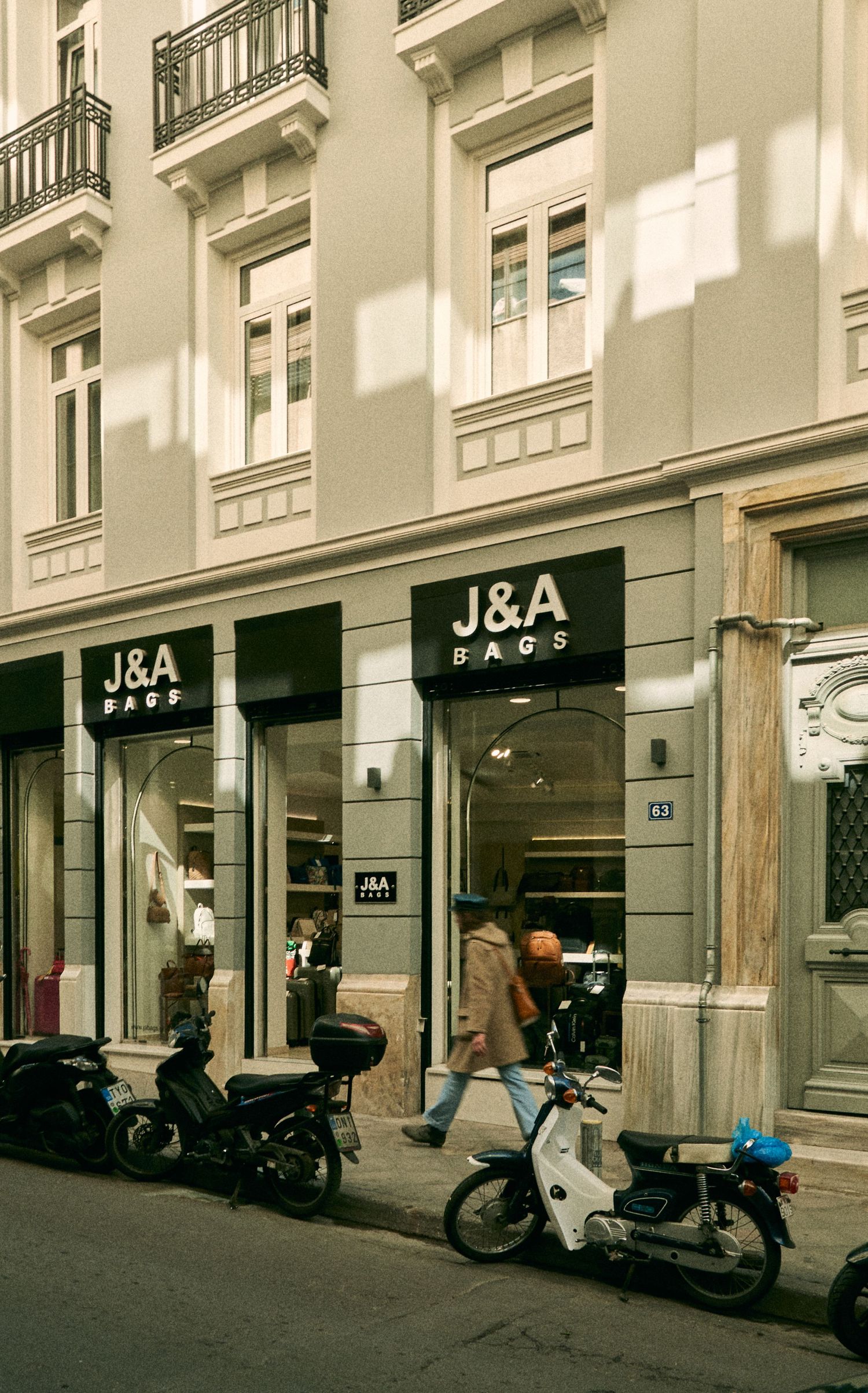 Storefront of J&A bags shop with motorcycles parked on a European city street.