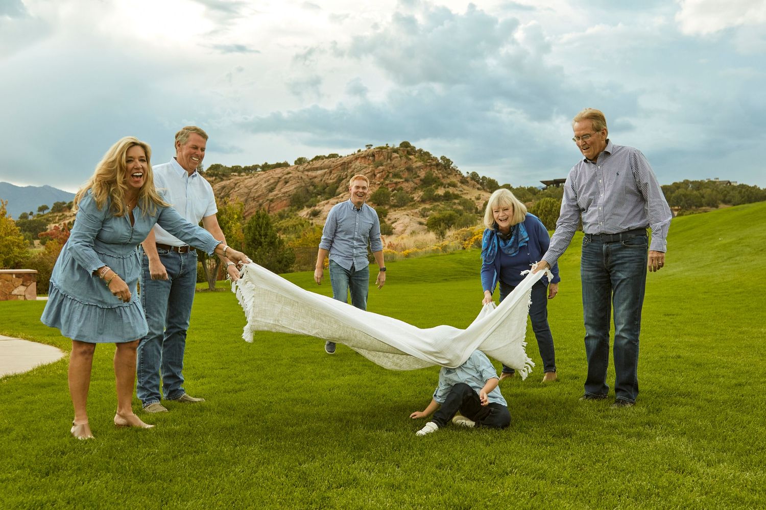 Family playing with blanket on grassy hill with mountains in background.