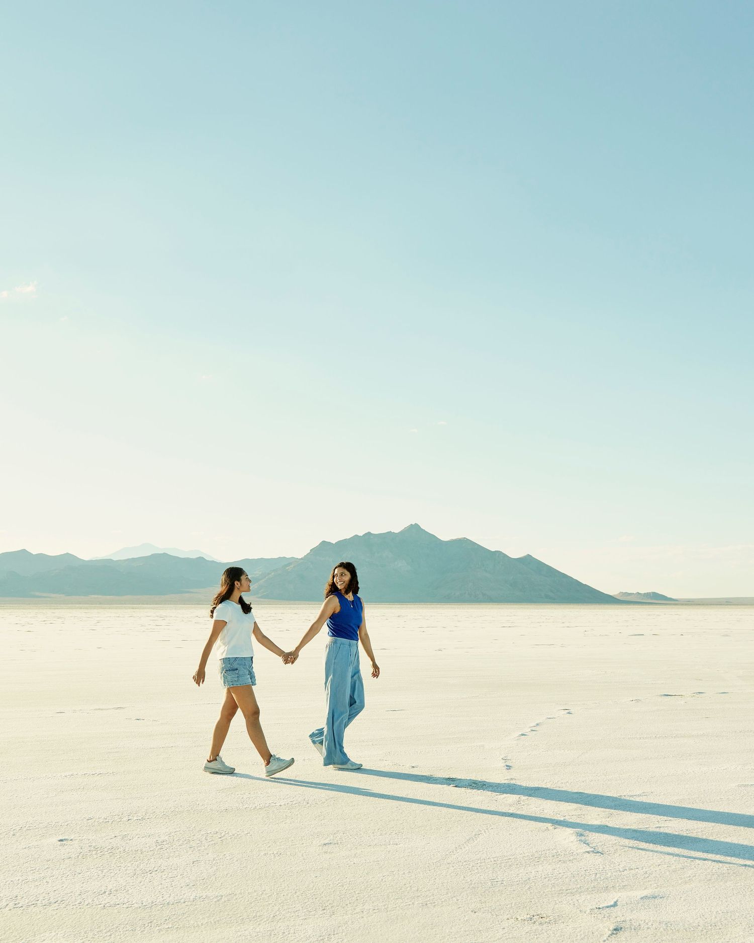 Two people walking hand in hand across white salt flats with mountains in distance.