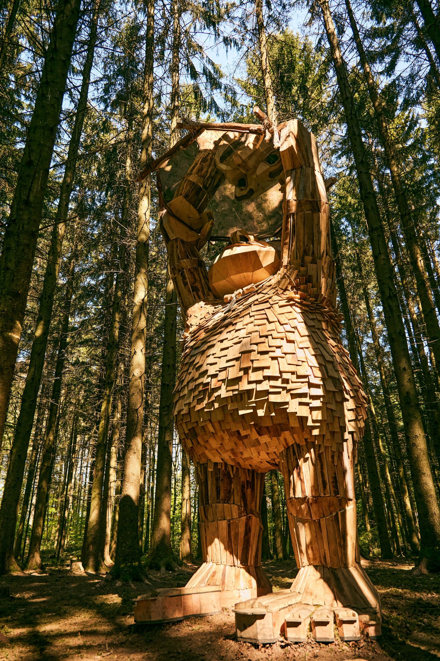 Giant wooden owl sculpture with intricate shingle details perched among tall evergreen trees in a forest.