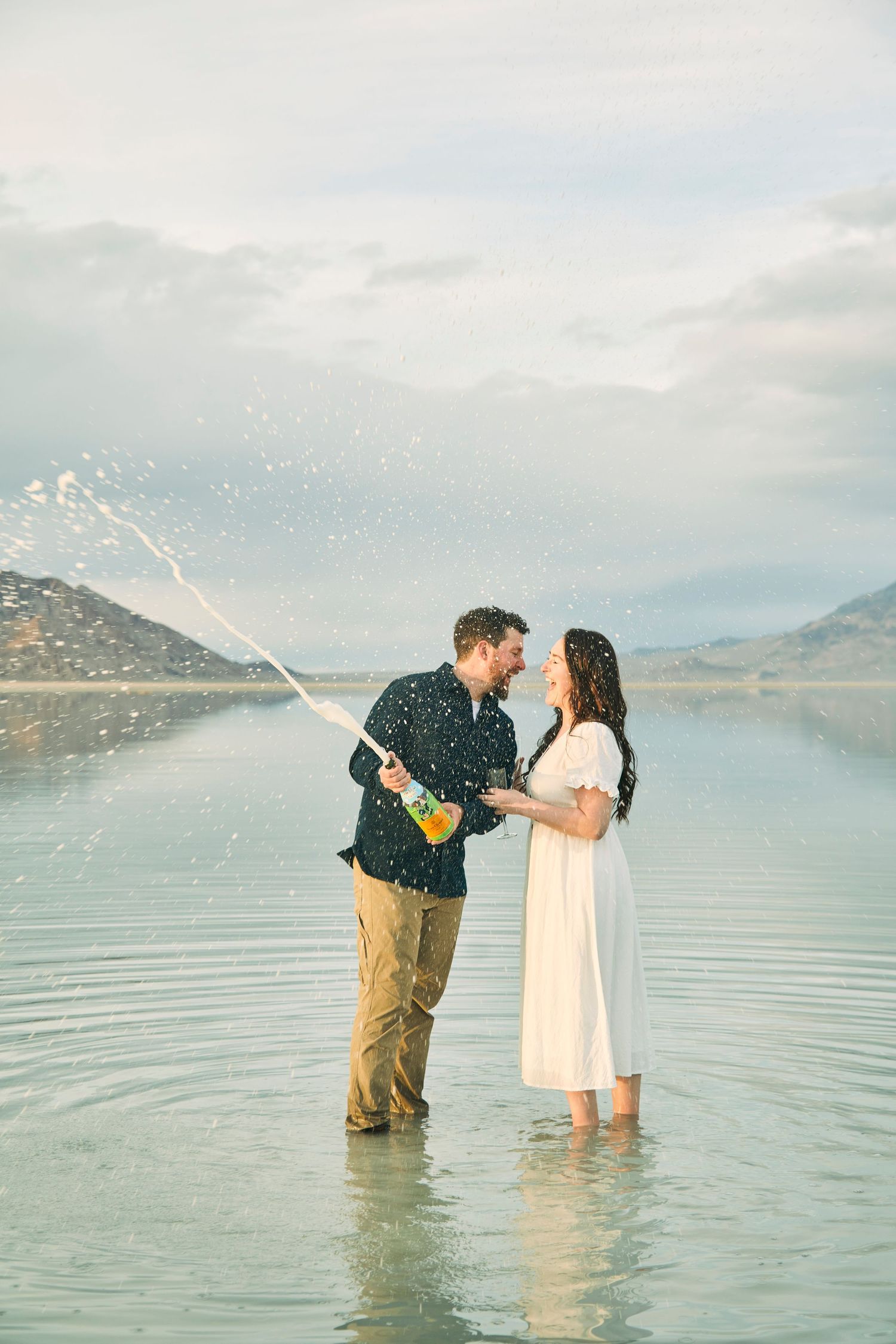 Couple celebrating with champagne while standing in shallow lake water with mountains in background.