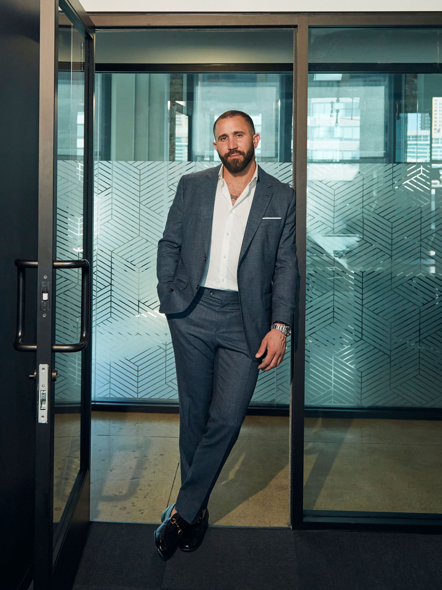 Business professional in modern office leans against glass wall wearing charcoal suit and white shirt.