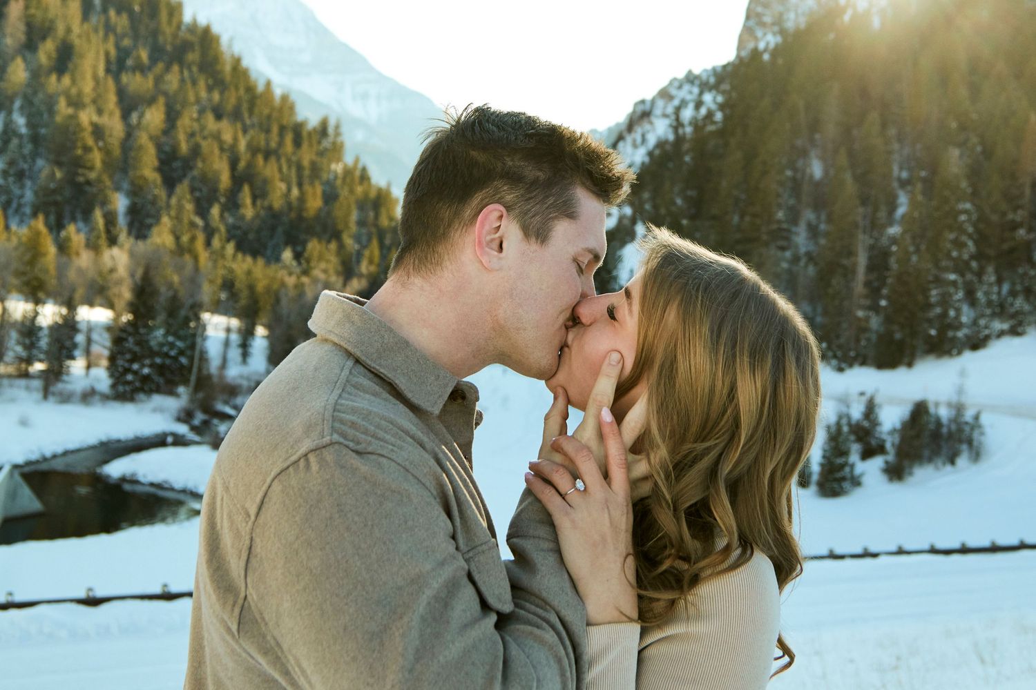 Couple sharing a romantic kiss in a snowy mountain setting at sunset.