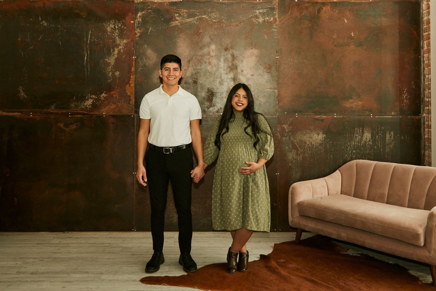 Couple poses against rustic industrial wall with vintage sofa in studio setting.