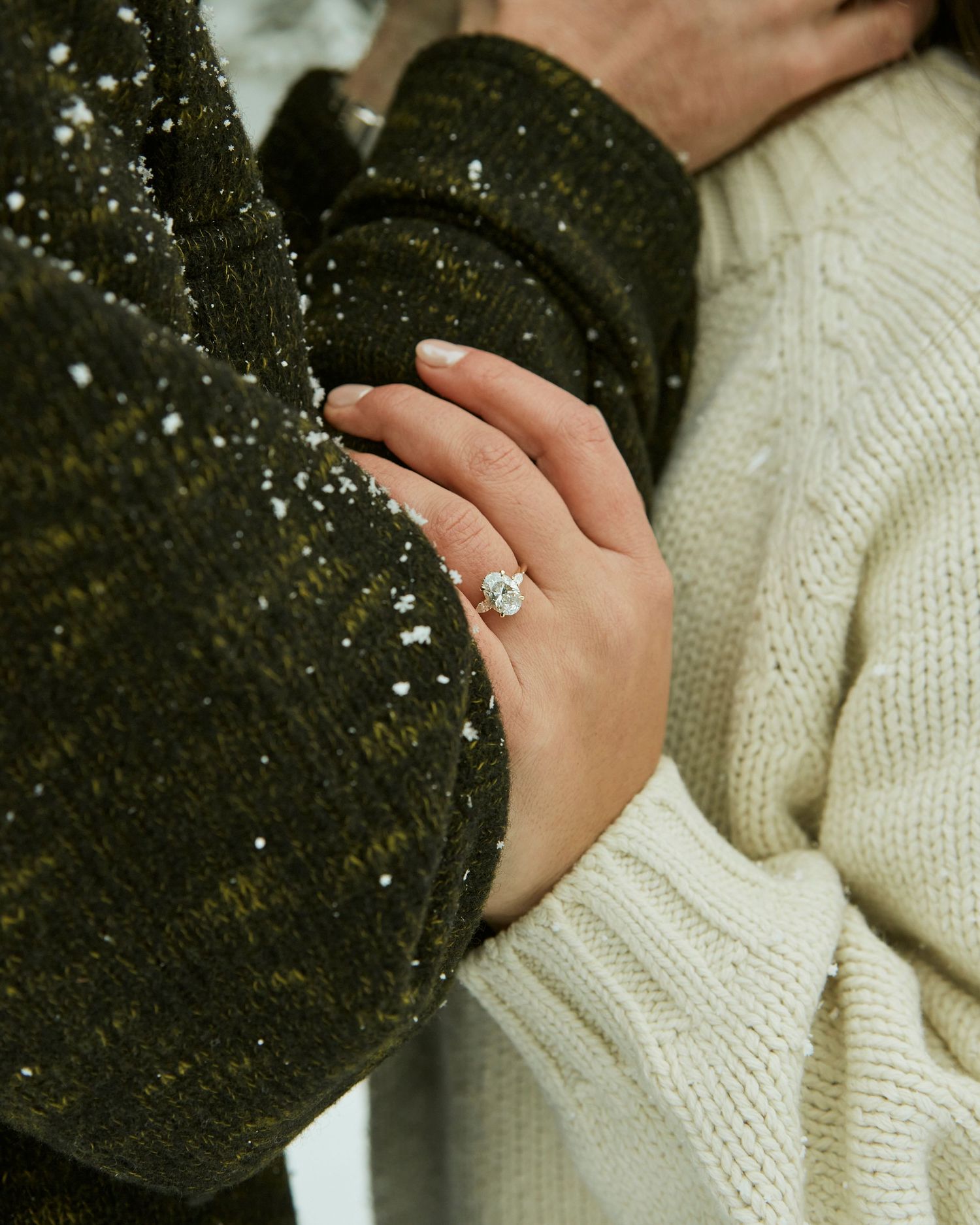 Close-up of an engagement ring on hand with snowflakes against cozy winter sweaters.